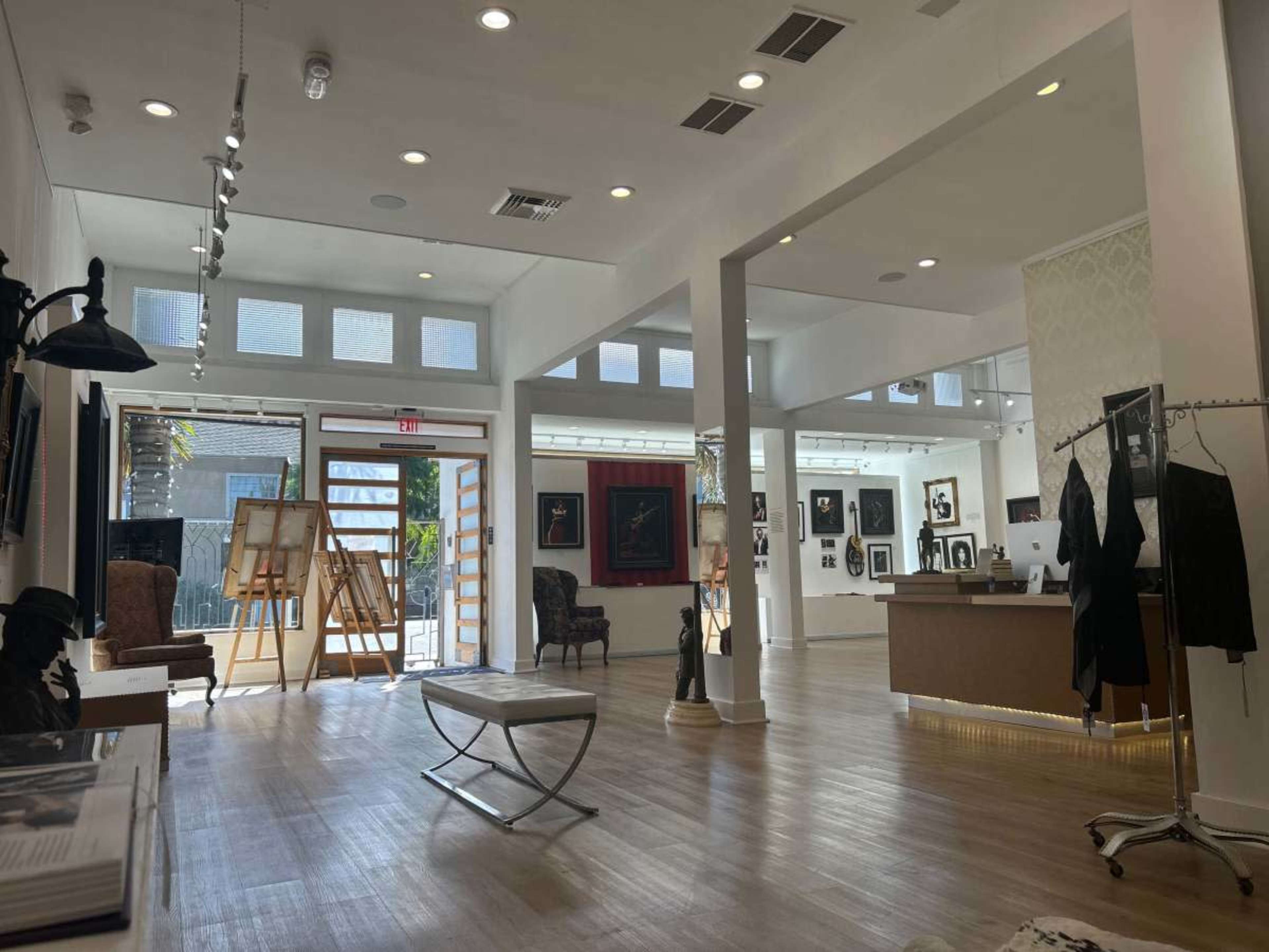 The image shows a spacious art gallery featuring wooden flooring, diverse artwork on the walls, and a reception area with seating.