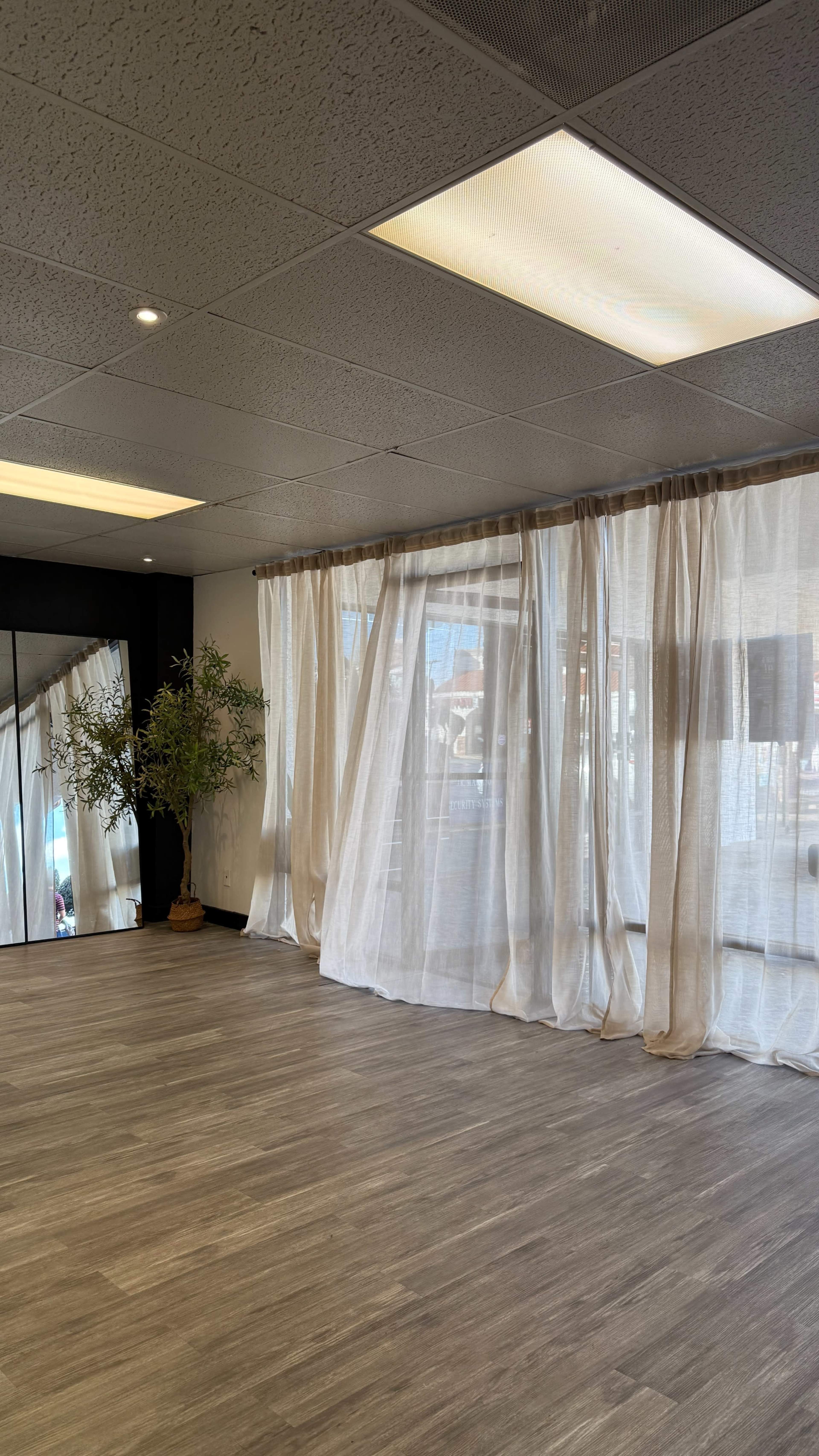 The image shows a spacious room with wooden flooring, large windows covered by sheer curtains, a potted plant in the corner, and a mirror on one wall.
