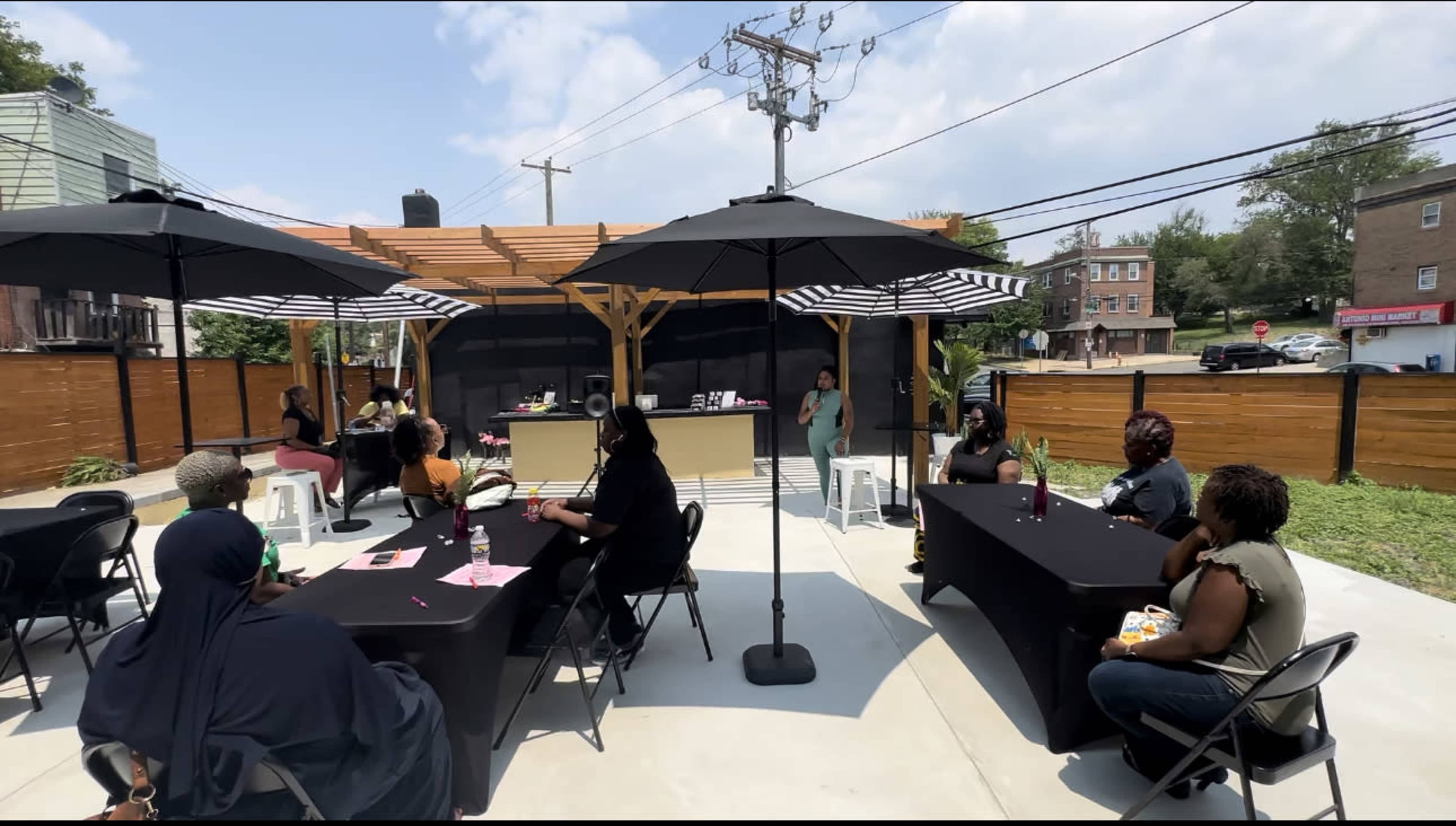 A group of people sits at tables under large umbrellas in an outdoor gathering space, while a person stands at the front speaking.