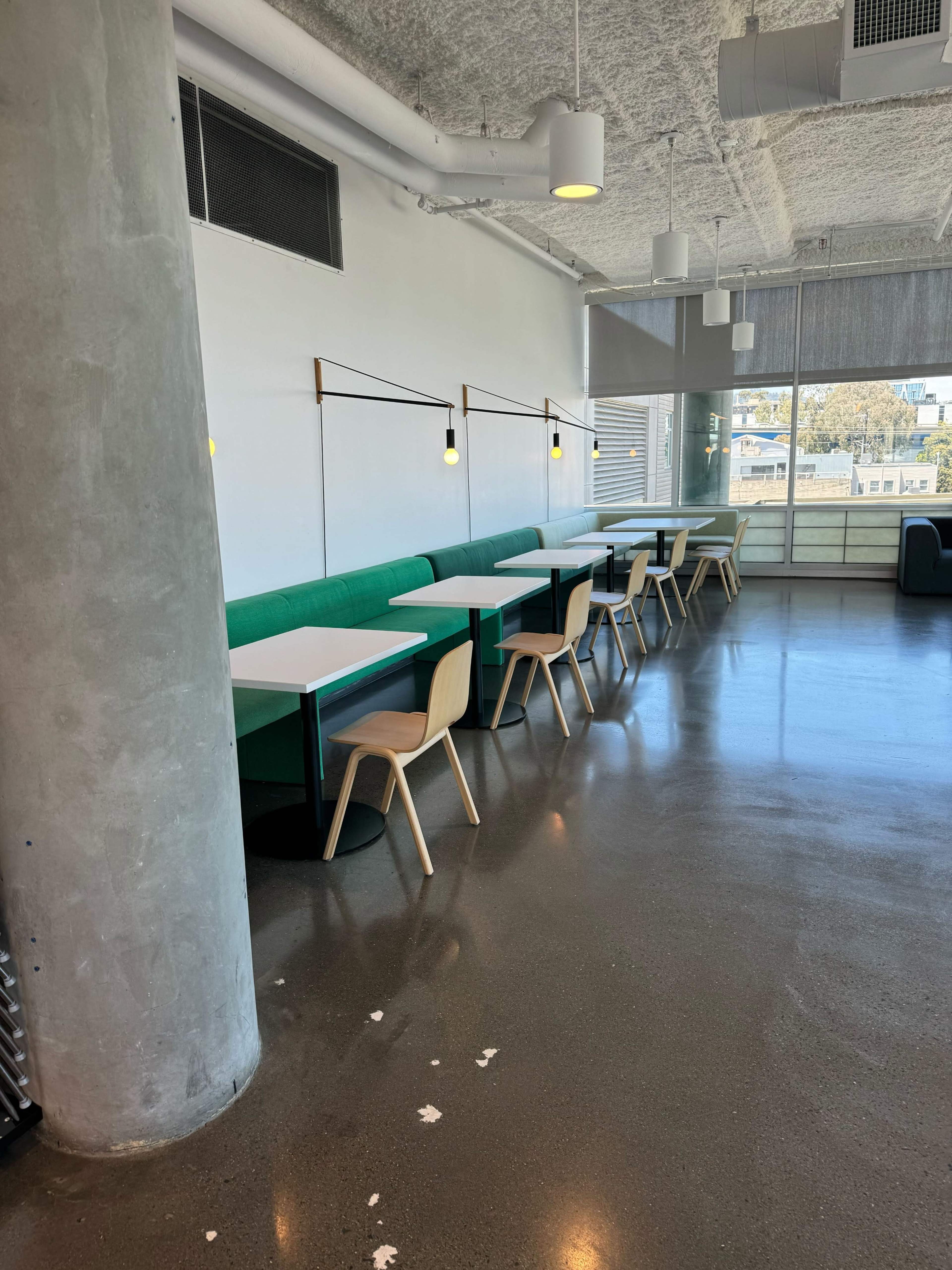 A modern interior space with green seating along the walls and white tables supported by wooden chairs, under bright overhead lighting.