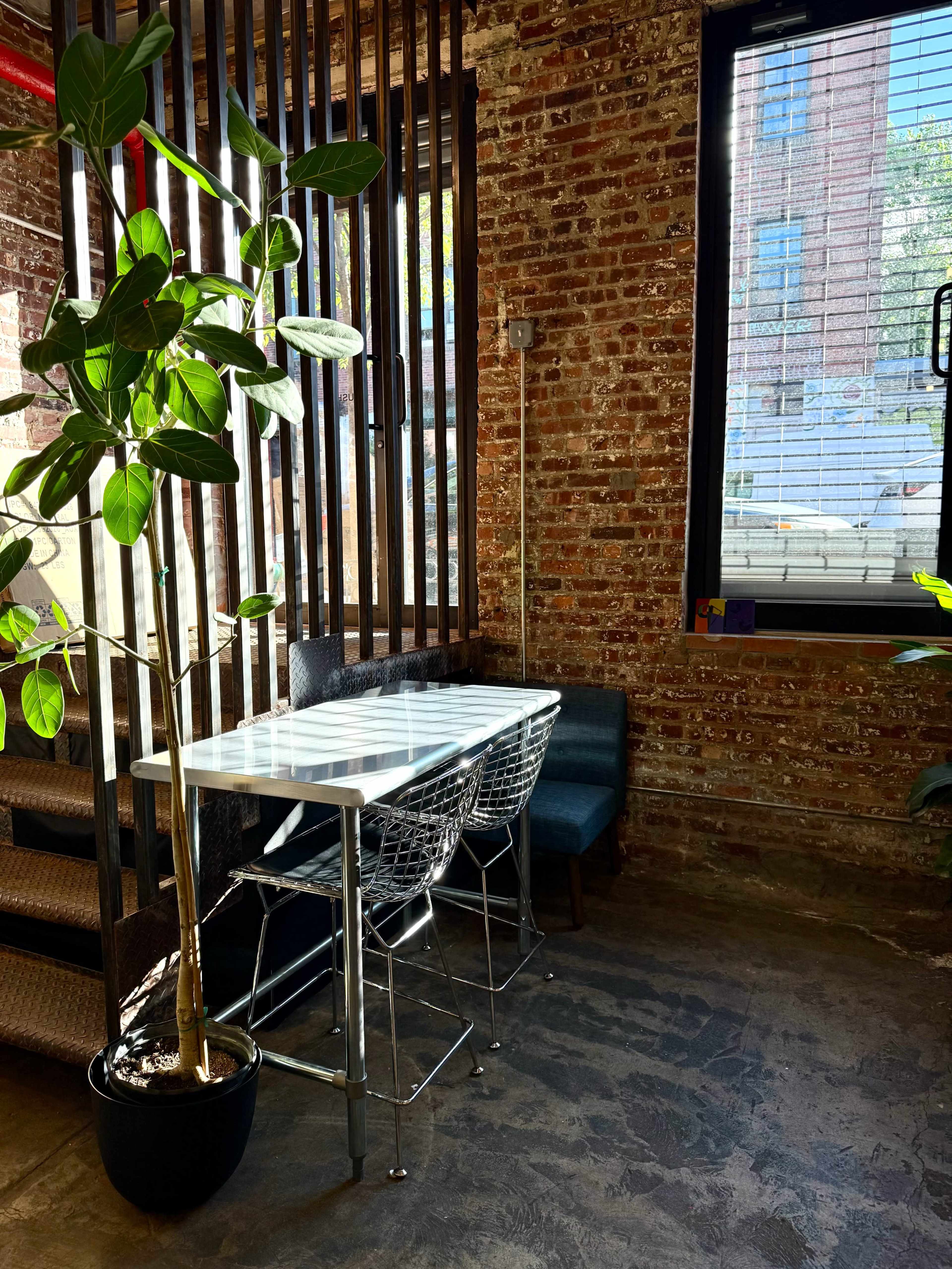 A modern, minimalist dining area features a glass table paired with wire chairs, surrounded by exposed brick walls and natural light from large windows.