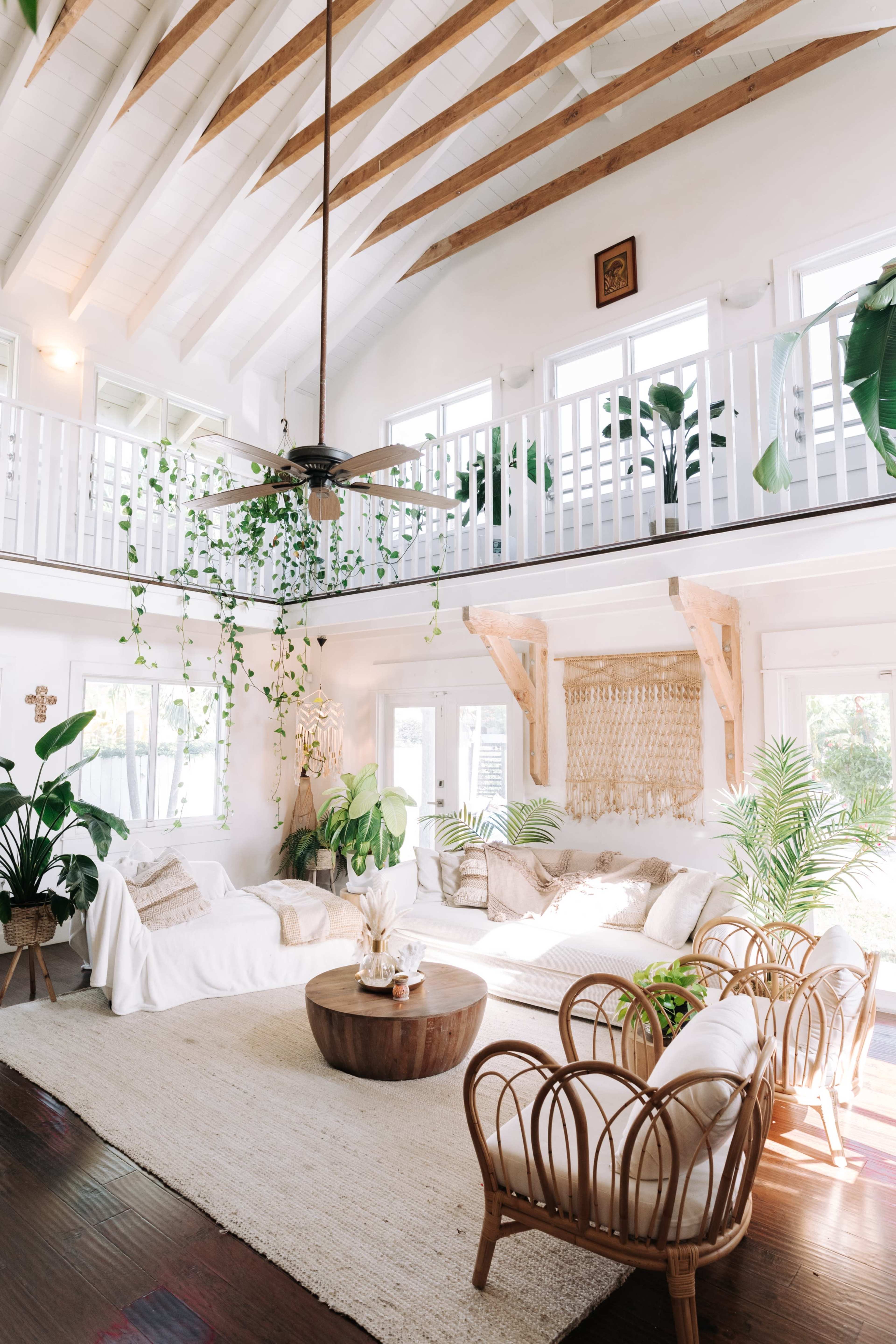 The image shows a bright, airy living room with high ceilings, wooden beams, and a large circular table surrounded by rattan chairs and various indoor plants.