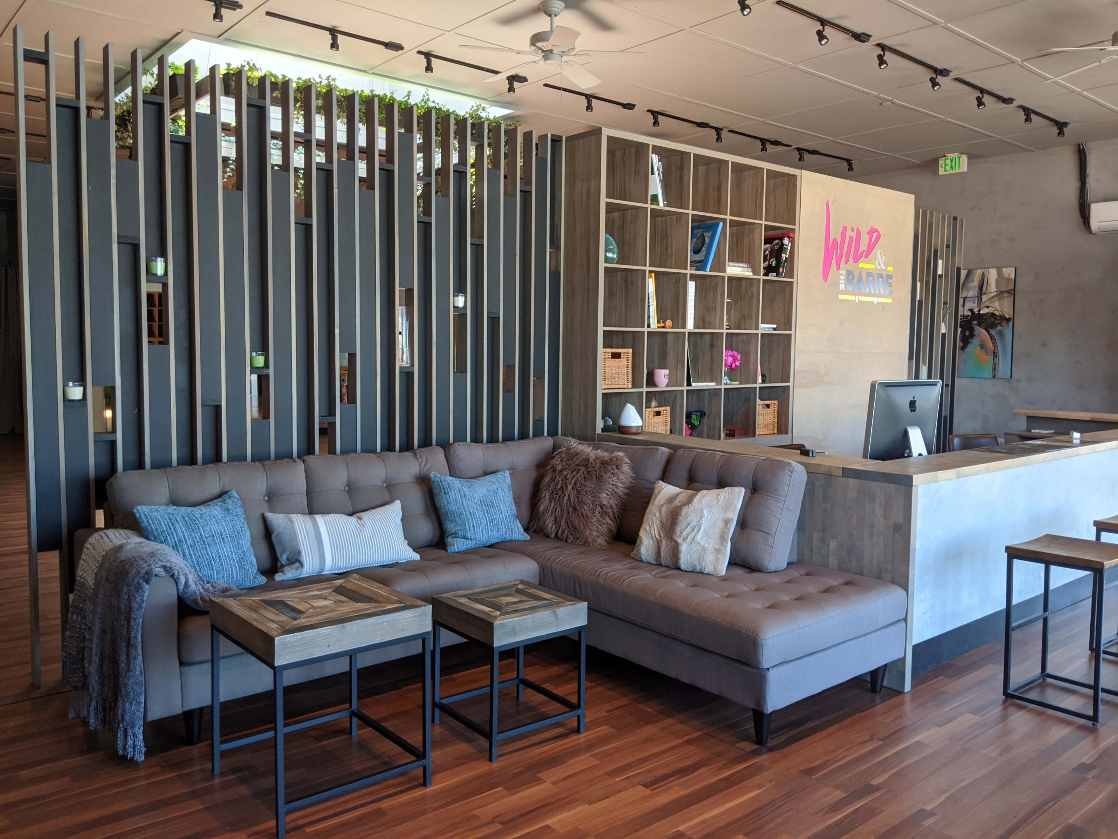 A modern reception area featuring a sectional sofa, wooden coffee tables, and a front desk against a backdrop of vertical wooden slats and shelves.