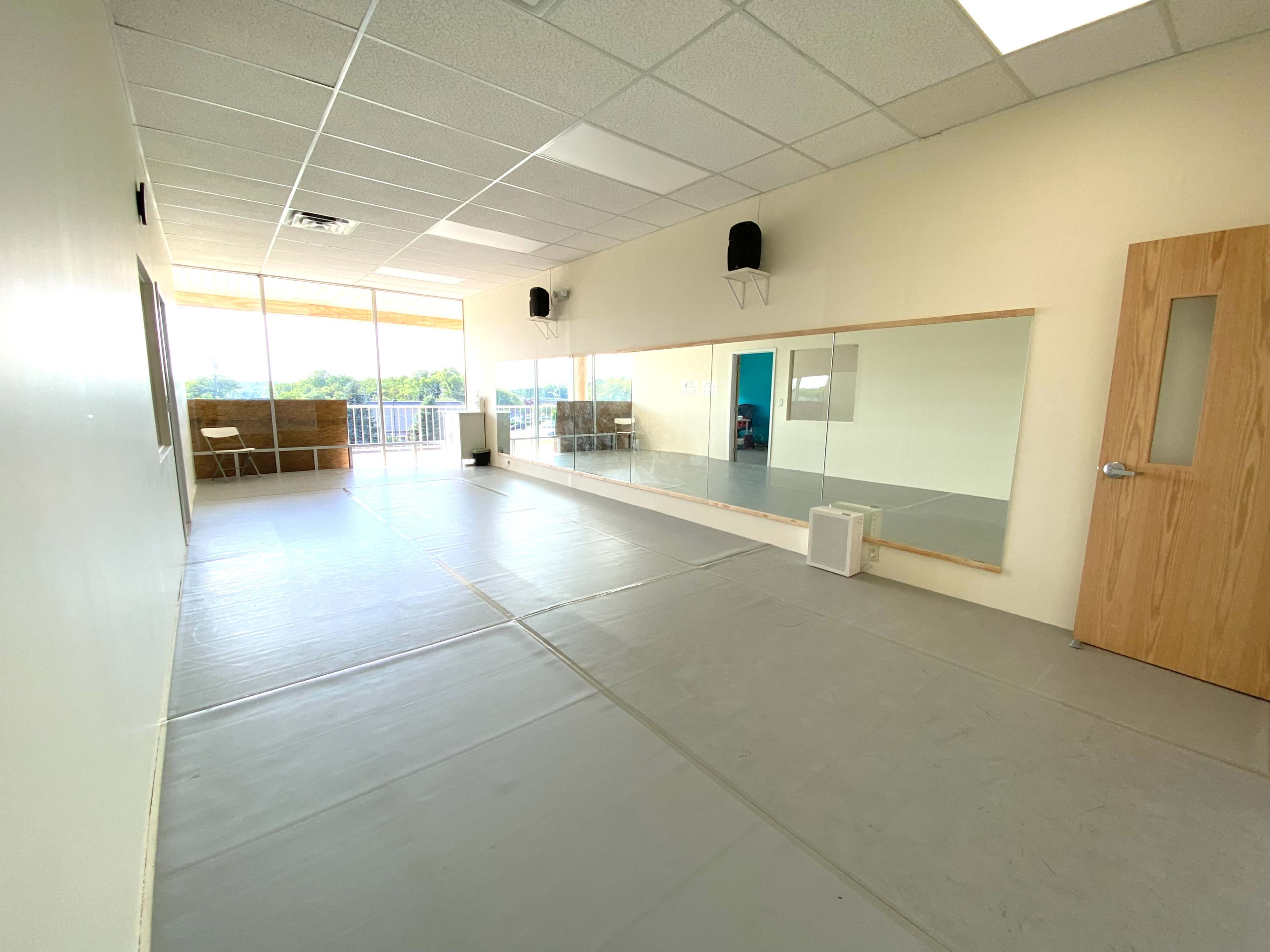 An empty dance studio with gray flooring, large mirrors, and natural light coming through a window.