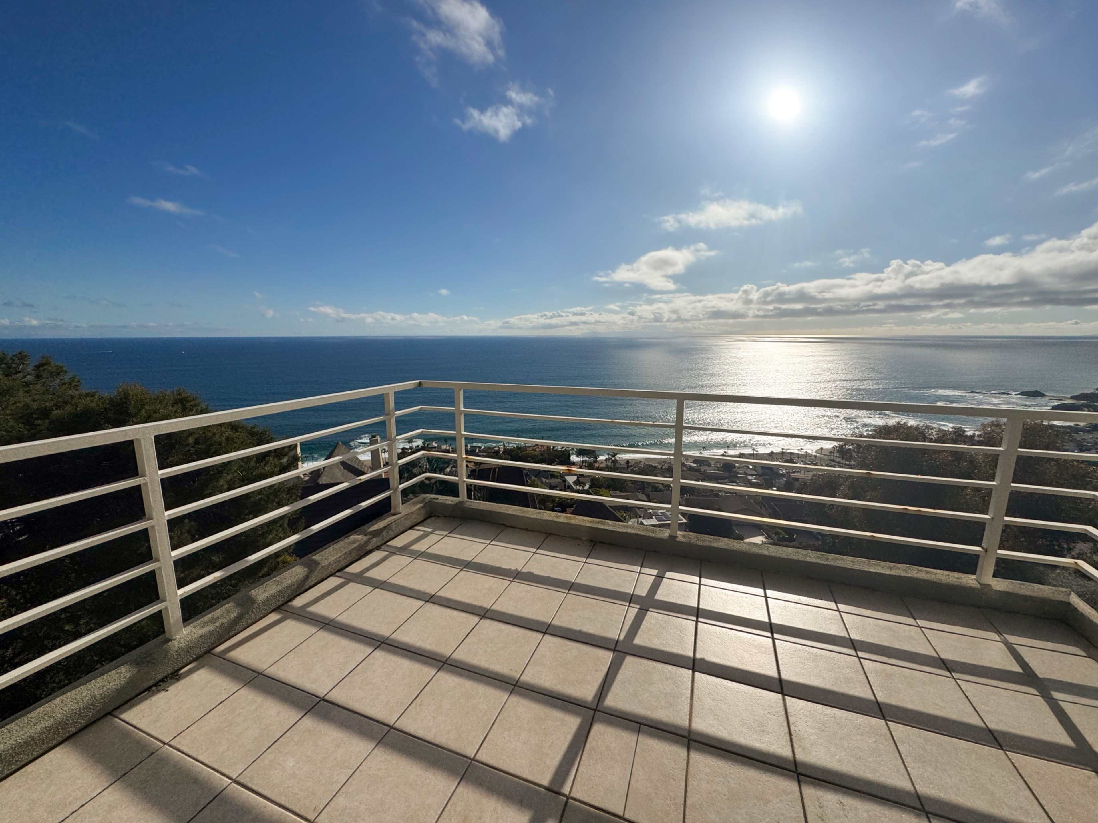 A spacious balcony overlooks the ocean, framed by a clear blue sky and sunlight reflecting off the water.
