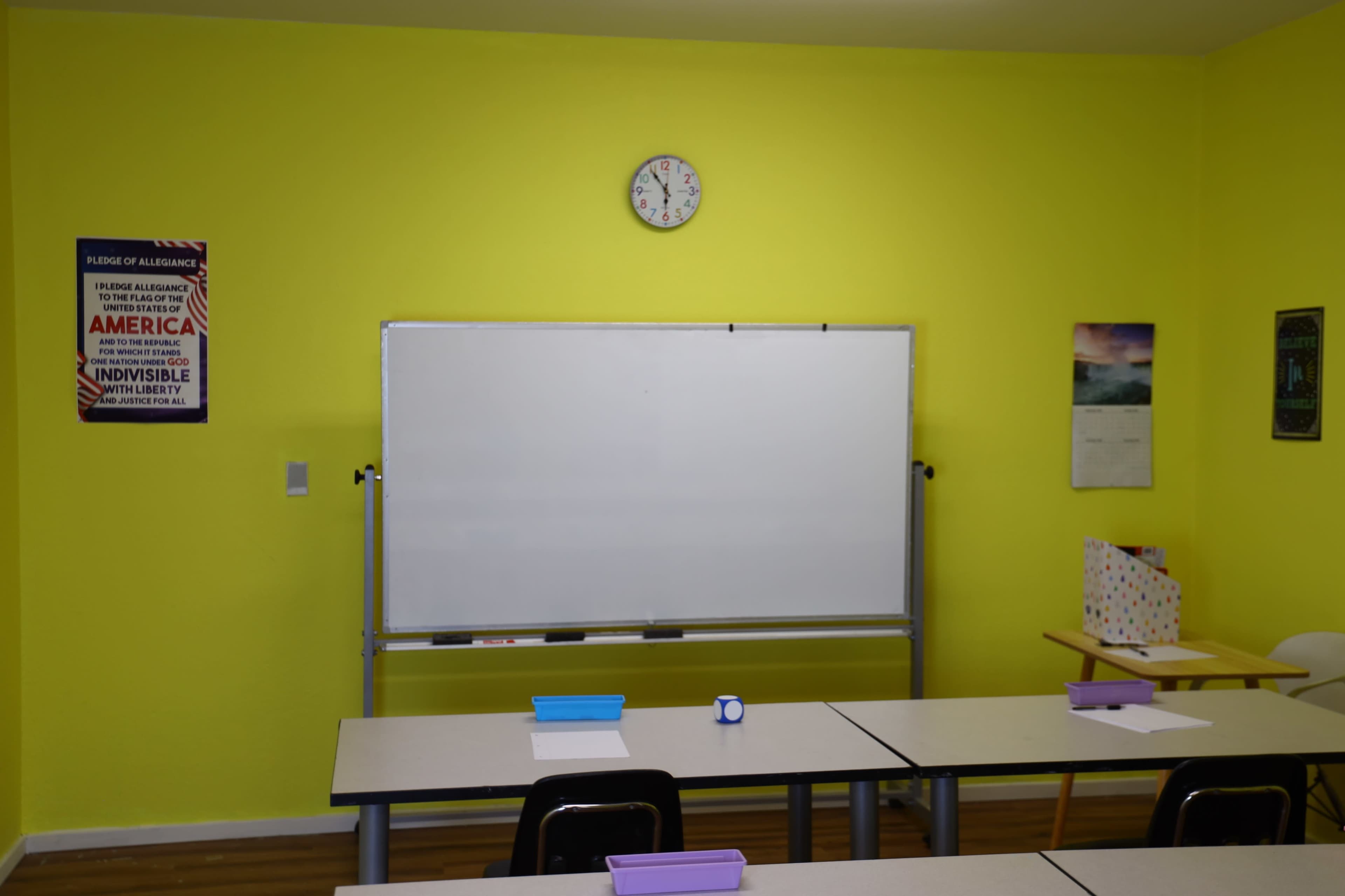 The image shows a brightly colored classroom with a yellow wall, a whiteboard on a stand, and several desks equipped with school supplies.