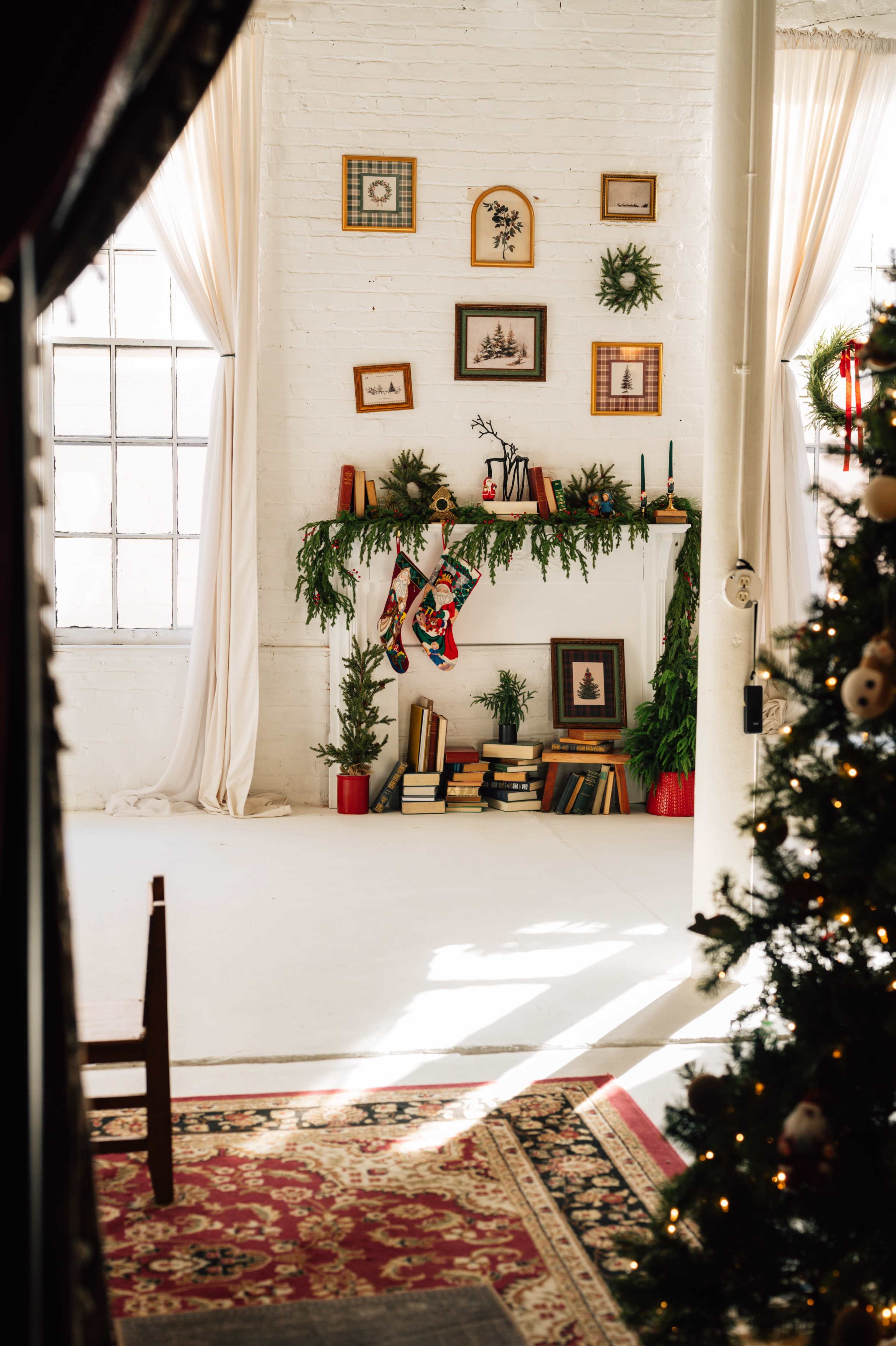 A bright room features a decorated mantel with festive greenery, stockings, and framed pictures, while a Christmas tree stands nearby.