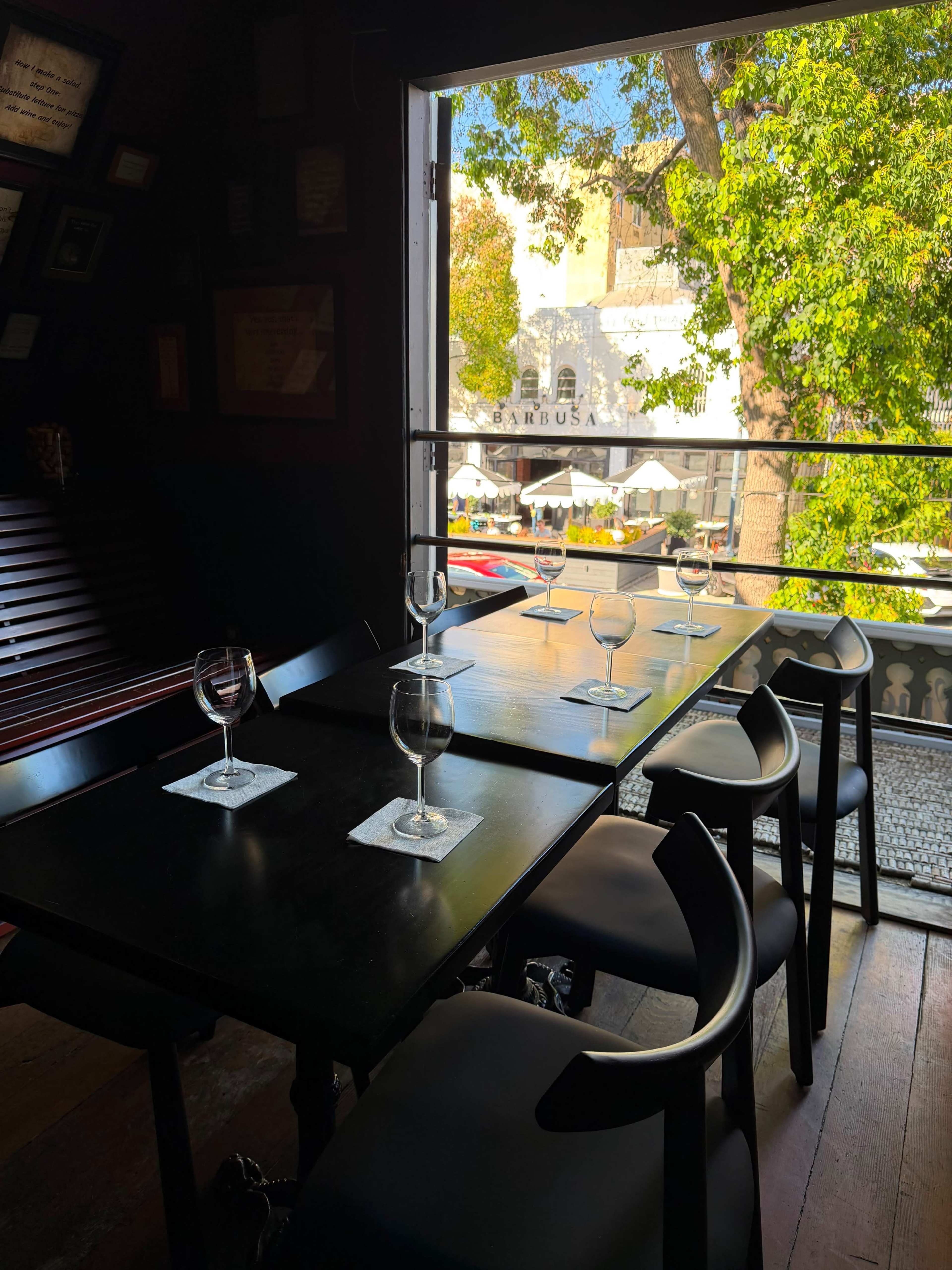 The image shows a table set with wine glasses in a restaurant, with a view of an outdoor dining area and trees through an open window.