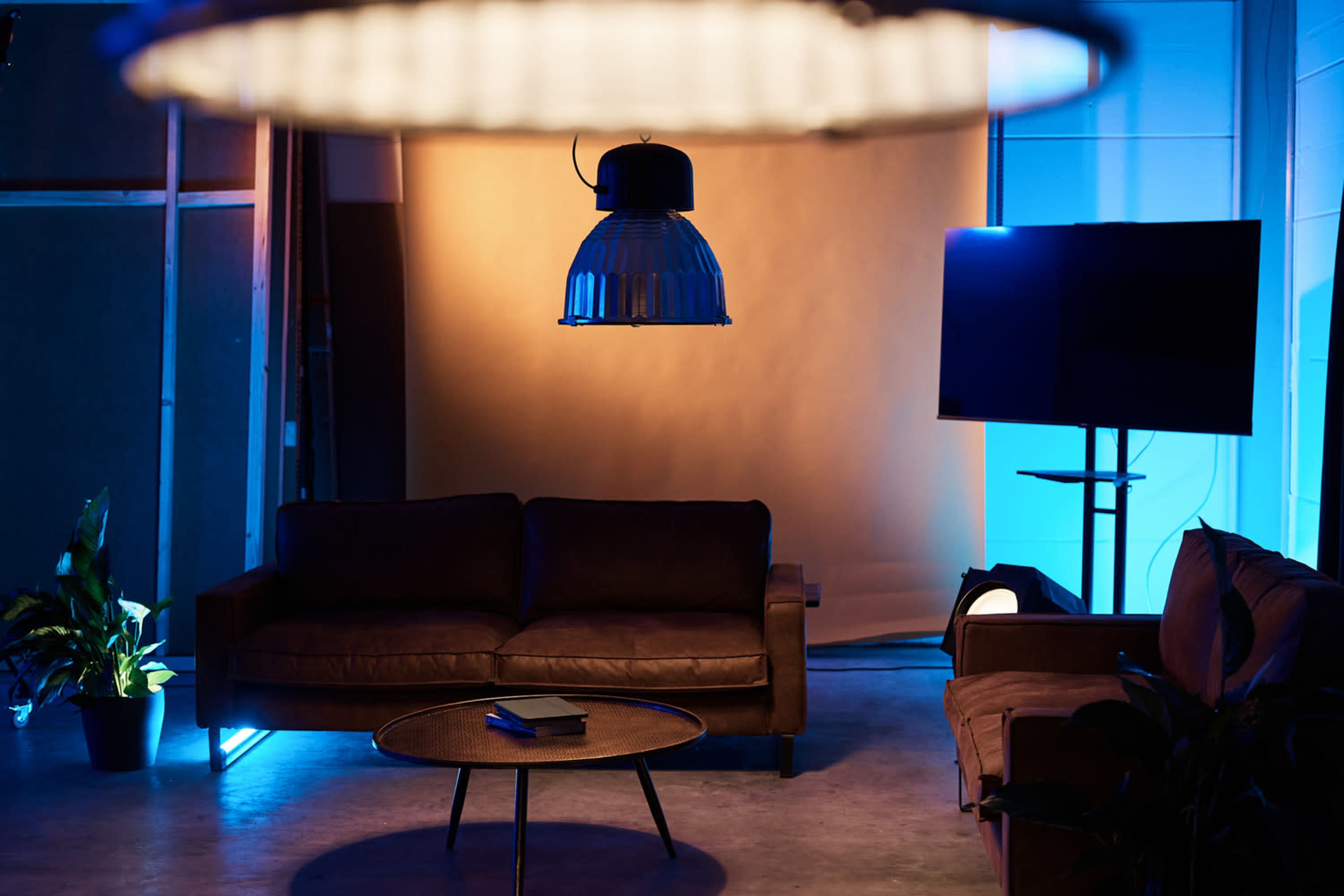 The image shows a modern lounge area with two sofas, a coffee table, a large pendant light, and a television on a stand, all set against a softly lit backdrop.