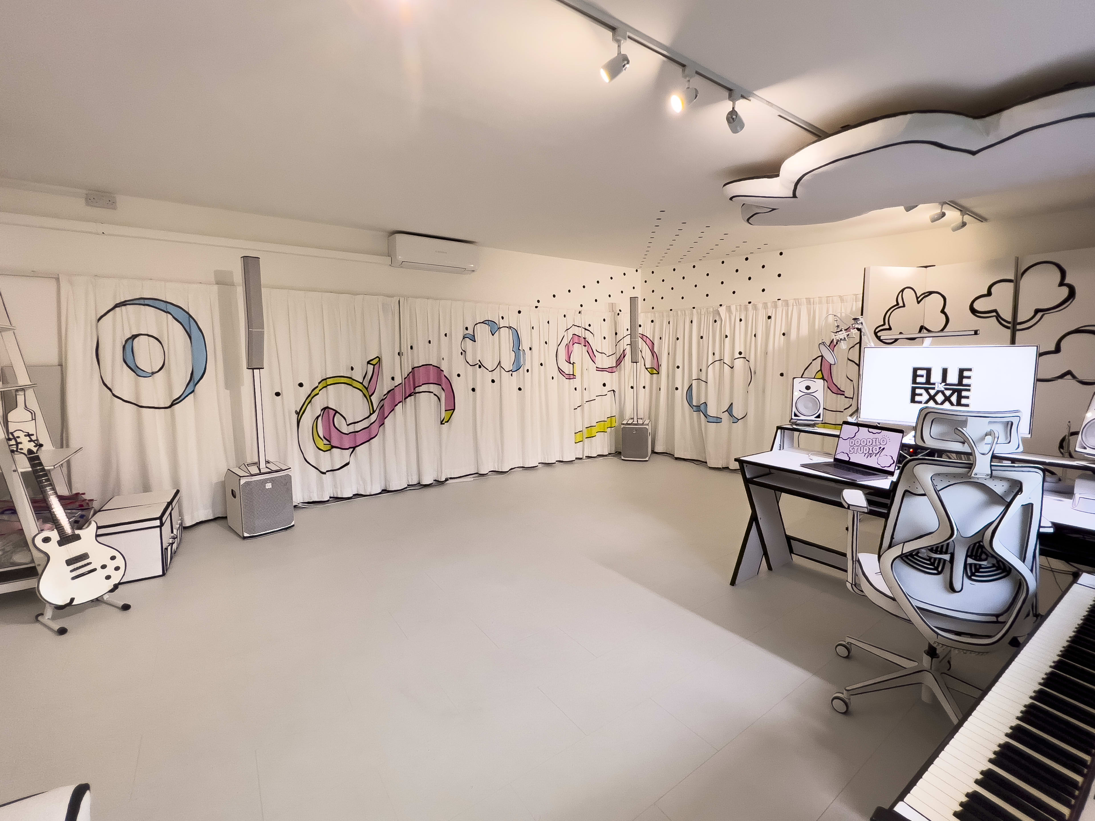 A modern studio space with white walls featuring colorful cartoon-style designs, a desk with computer equipment, and musical instruments against a light-colored floor.