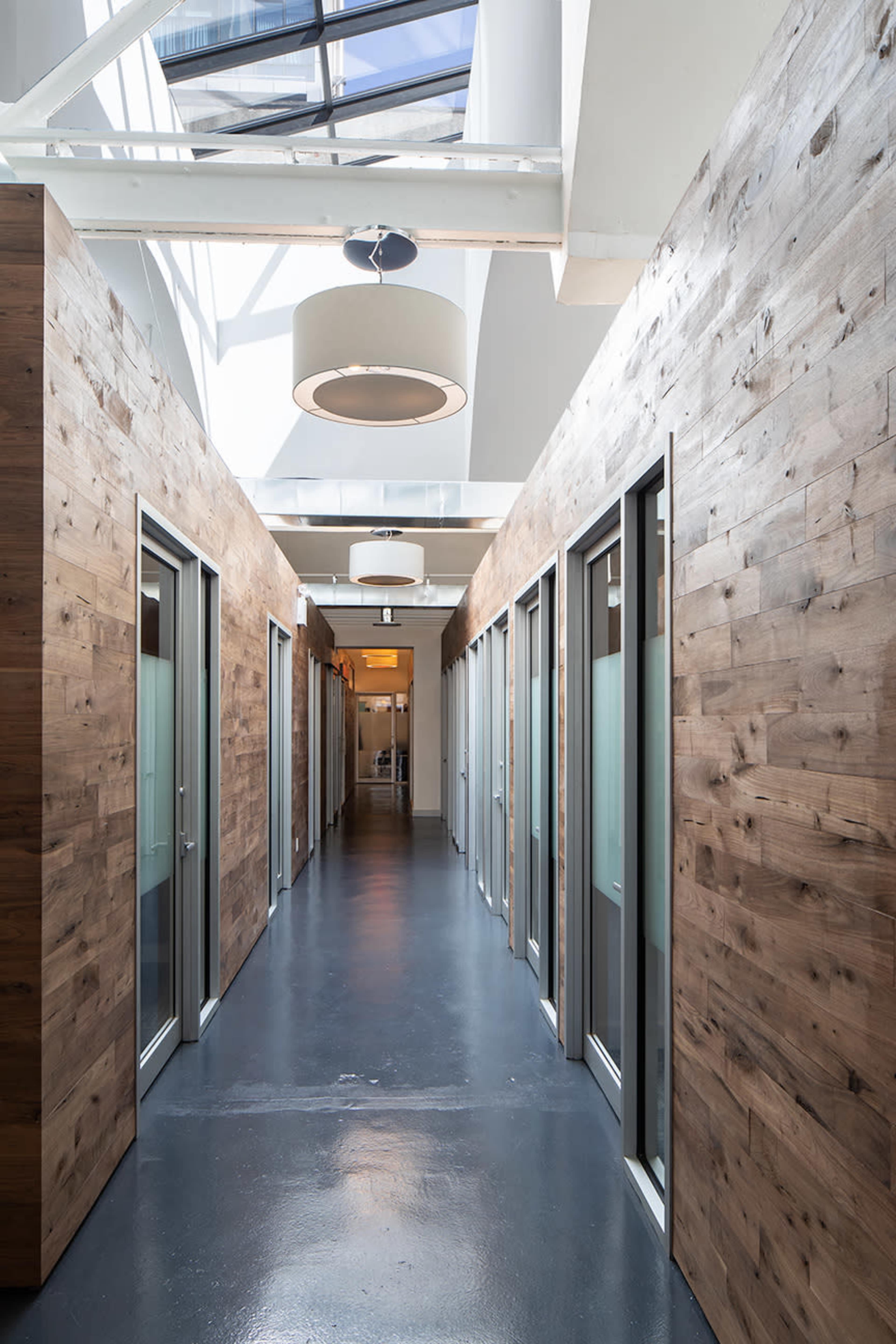 A modern hallway lined with wooden-paneled offices and overhead lighting, featuring a skylight that illuminates the space.