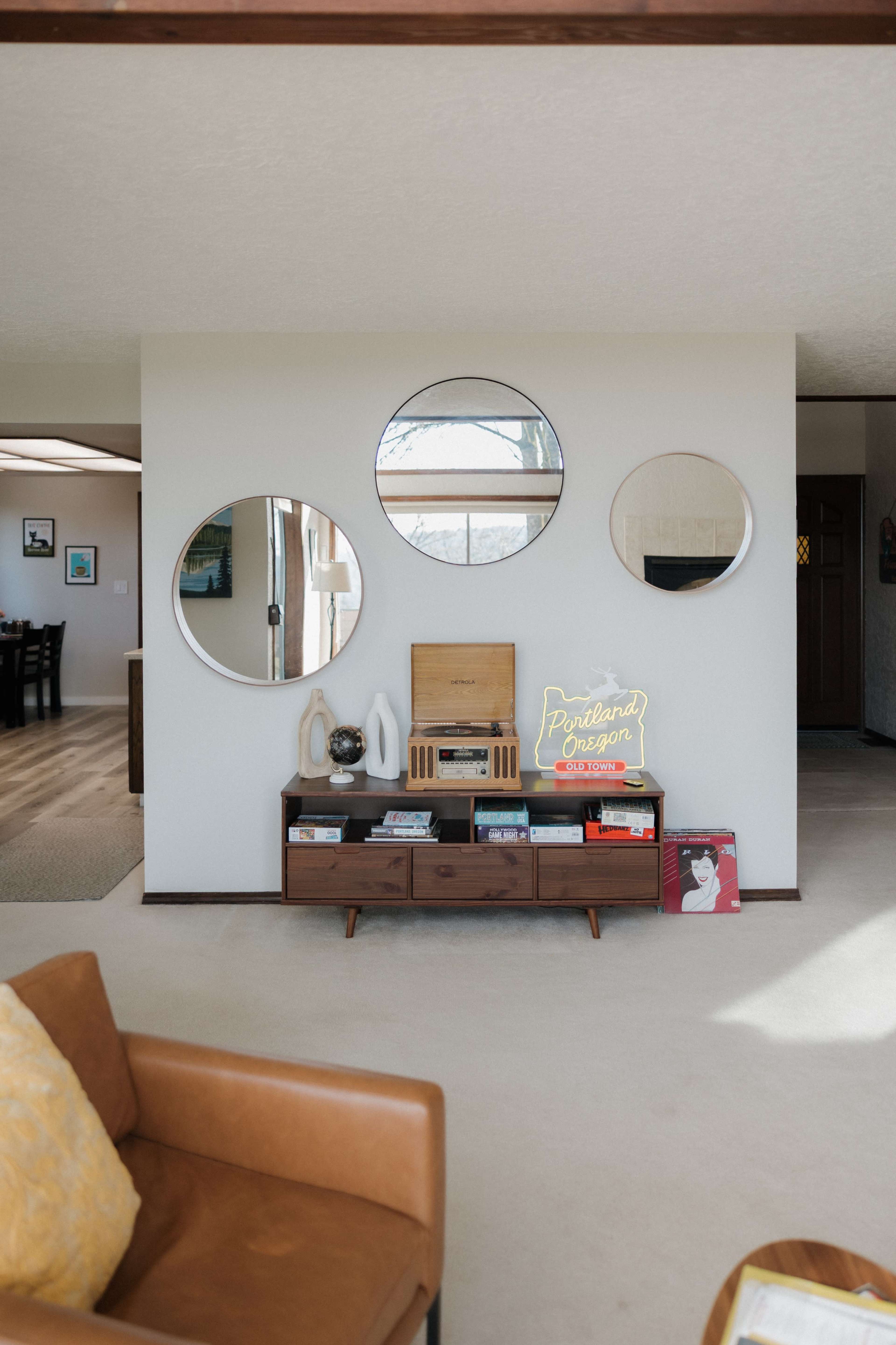 The image shows a modern living room with a wooden sideboard featuring a record player, a neon sign, and three round mirrors mounted on the wall above it.
