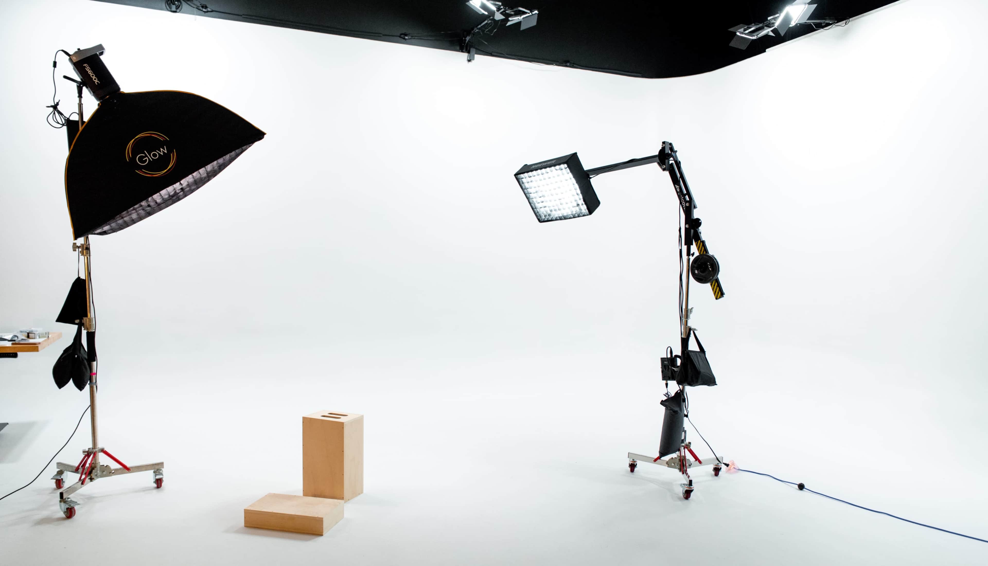 The image shows a photography studio with two lighting setups and a wooden block placed in the center of an empty white backdrop.