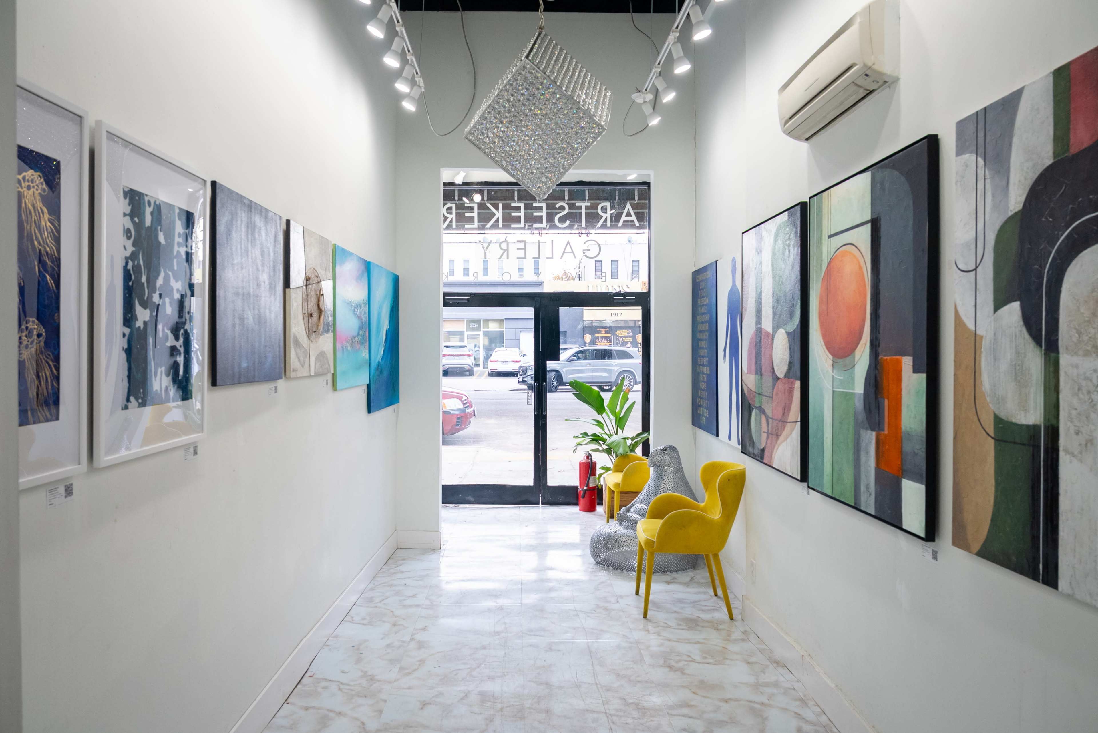 The image shows a narrow art gallery hall featuring several framed artworks on the walls, with a yellow chair and a potted plant positioned near the entrance.