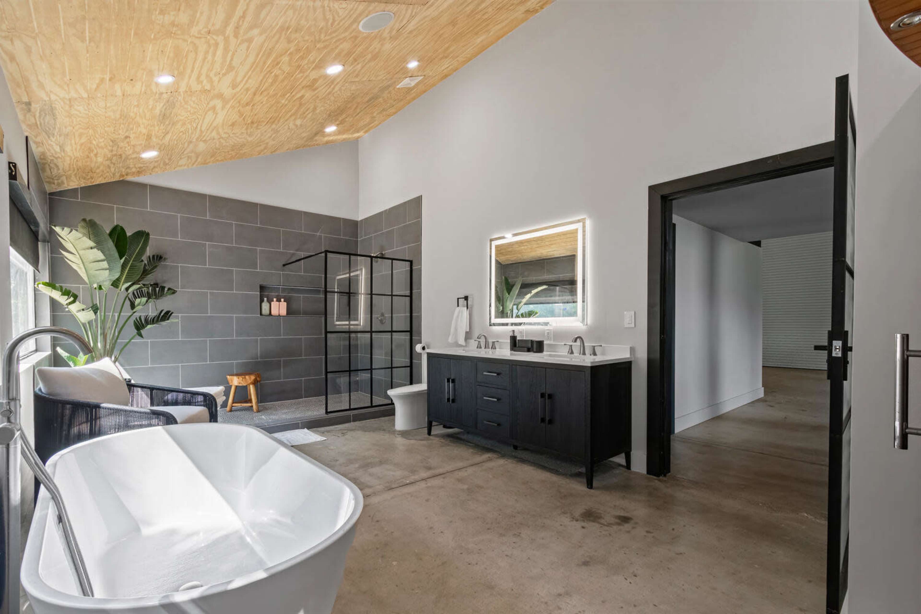 The image shows a modern bathroom featuring a freestanding tub, a sleek black double vanity with a mirror, a glass shower enclosure, and a minimalist design with large tiles and natural light.