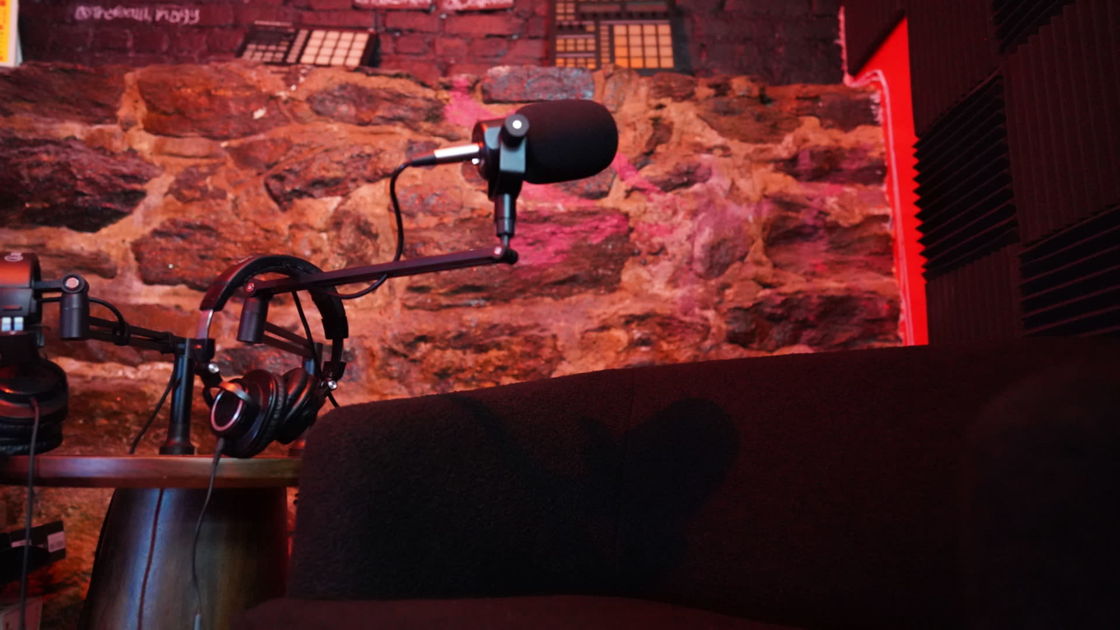 A microphone is mounted on a stand next to headphones, set against a textured stone wall illuminated with red lighting.