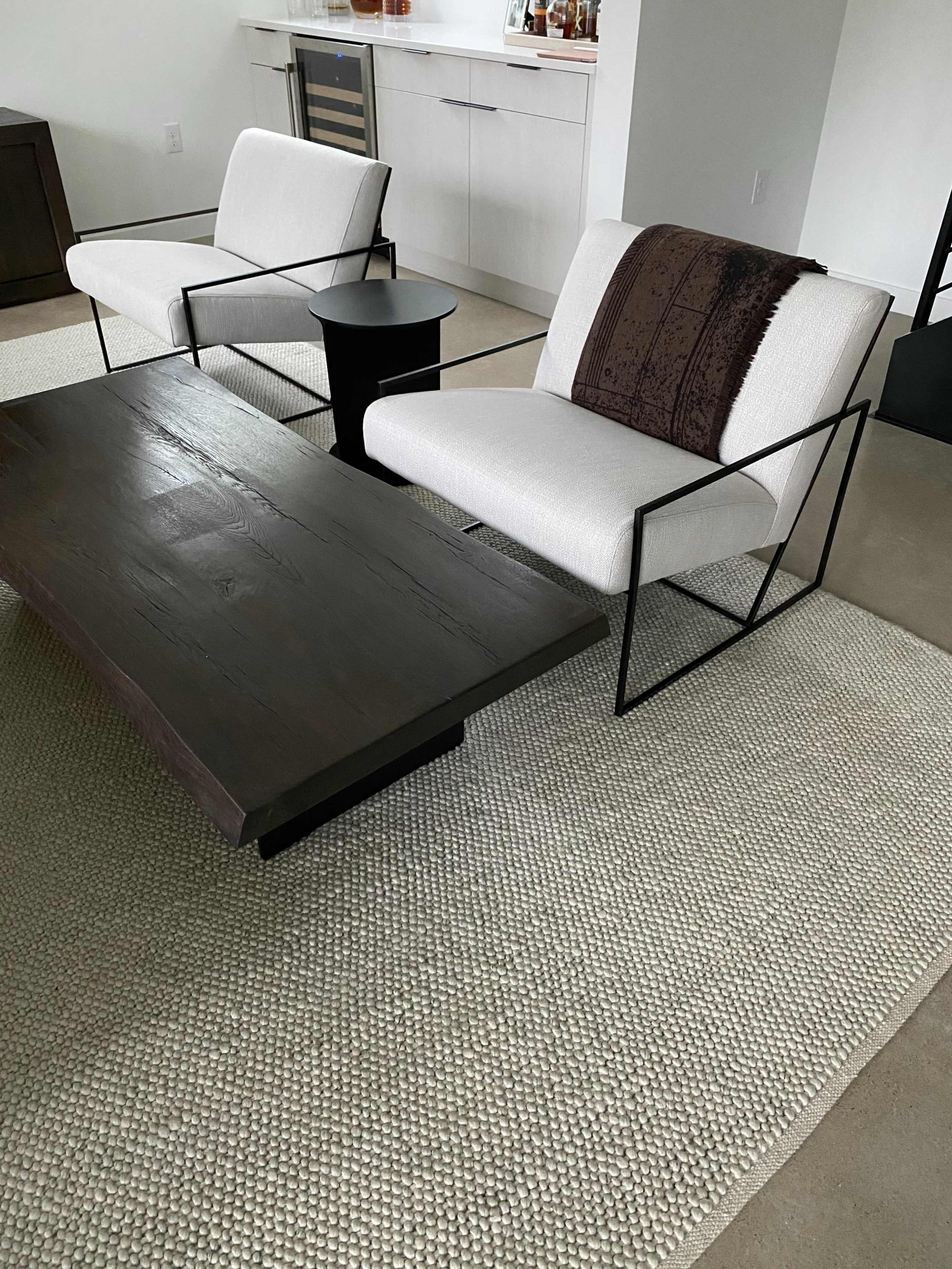 The image shows a cozy living area featuring two modern armchairs with a small side table and a large wooden coffee table on a textured area rug.