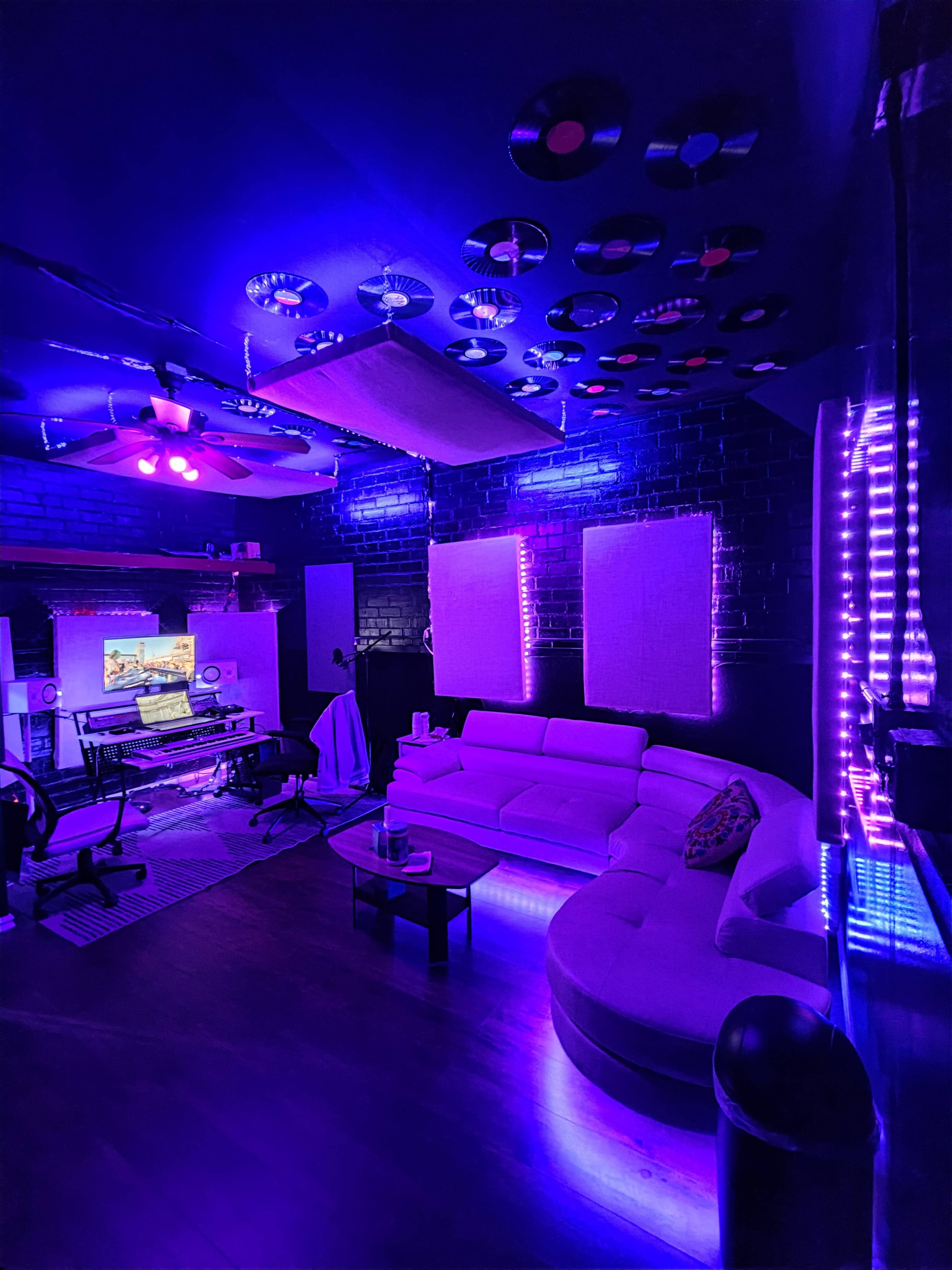 The image shows a modern music studio with a plush white sectional couch, purple LED lighting, and various music equipment arranged around the room.