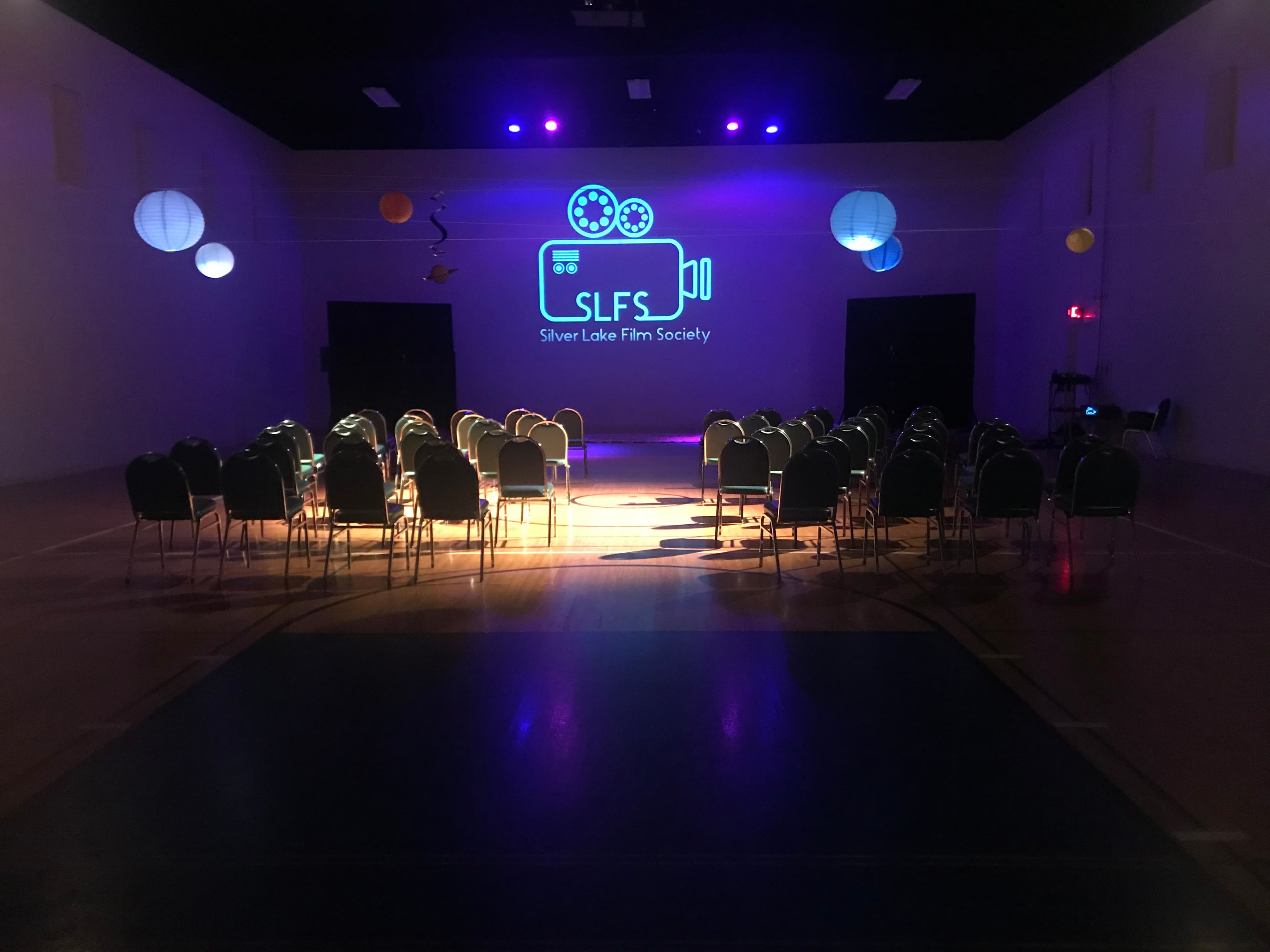 An empty auditorium set up for a Silver Lake Film Society event, with rows of chairs facing a screen displaying the society's logo and name, illuminated by purple lighting.