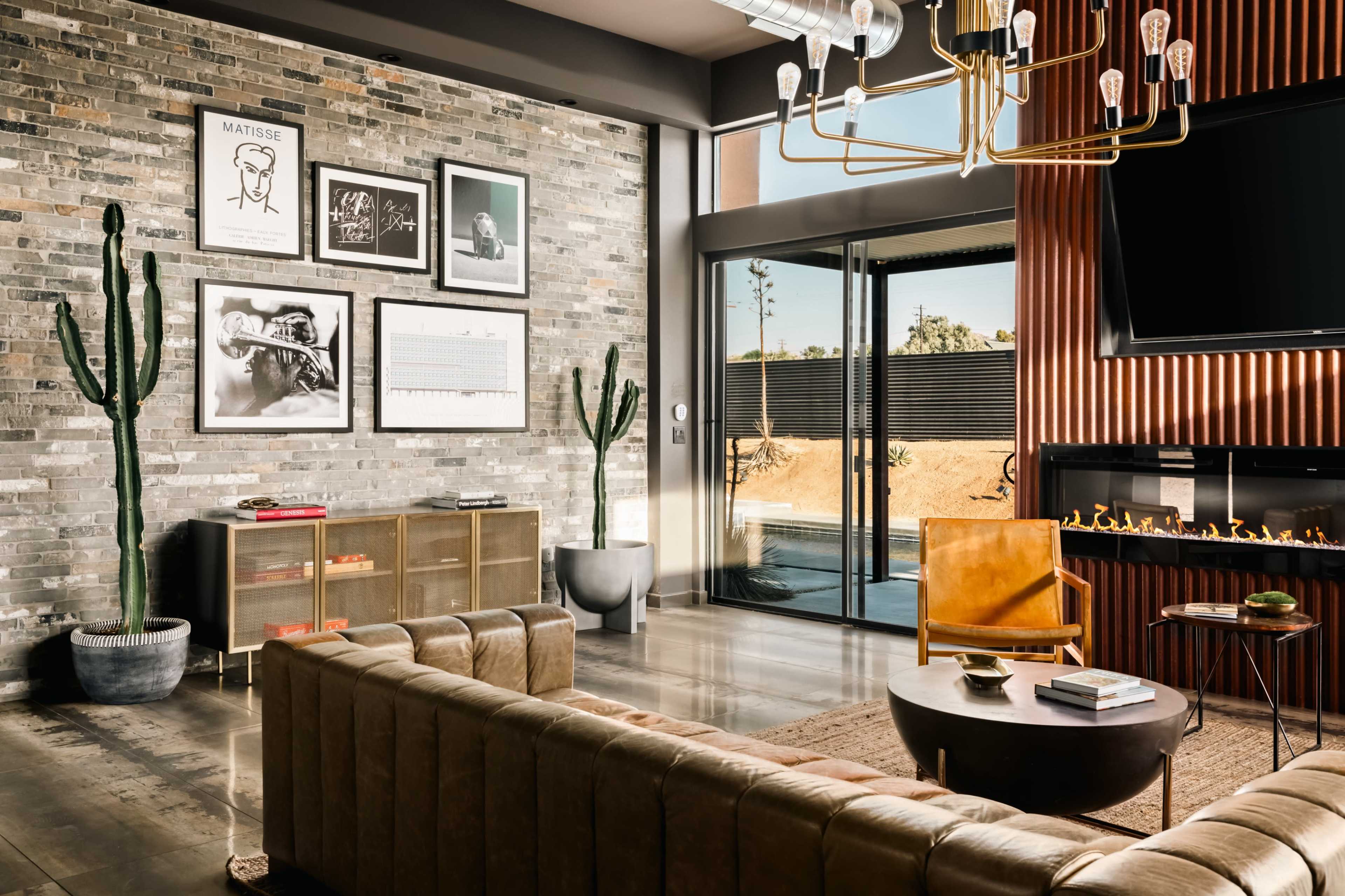 The living room features a modern design with a brick wall, framed artwork, cacti, a sectional sofa, and a view of an outdoor area through large glass doors.