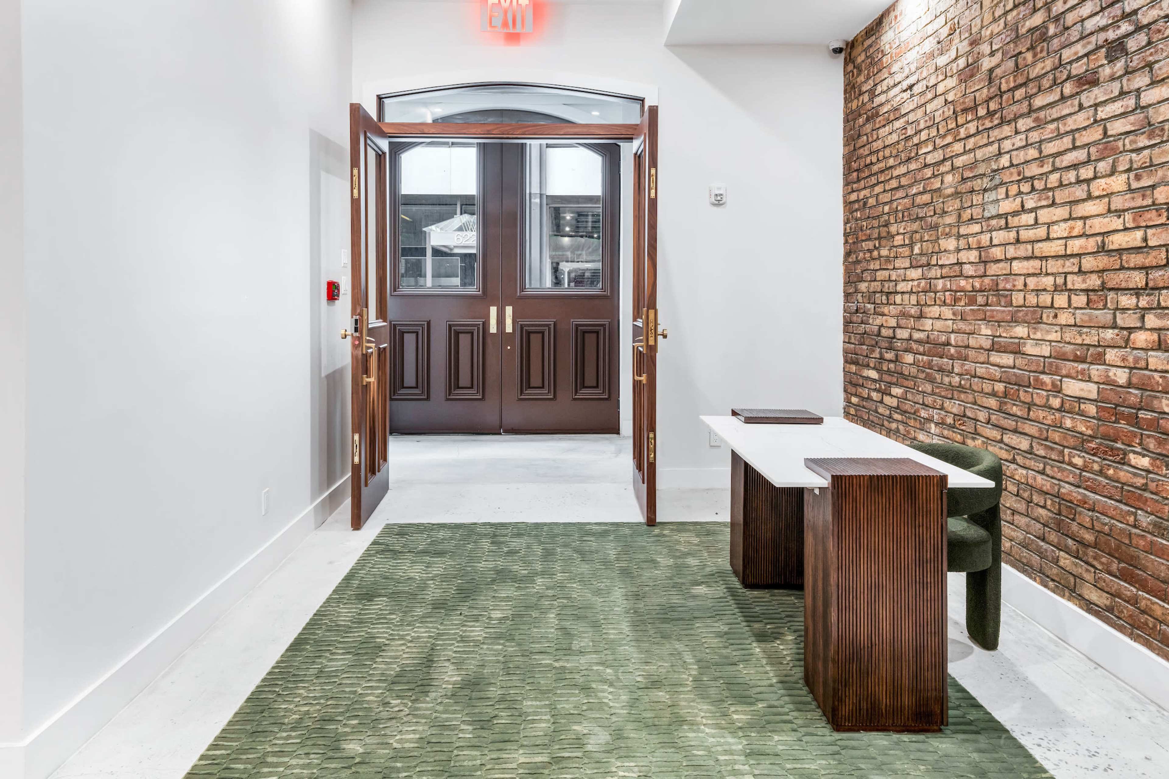 A hallway with an open entrance door, a textured brick wall on one side, and a small wooden reception desk next to a green carpet.