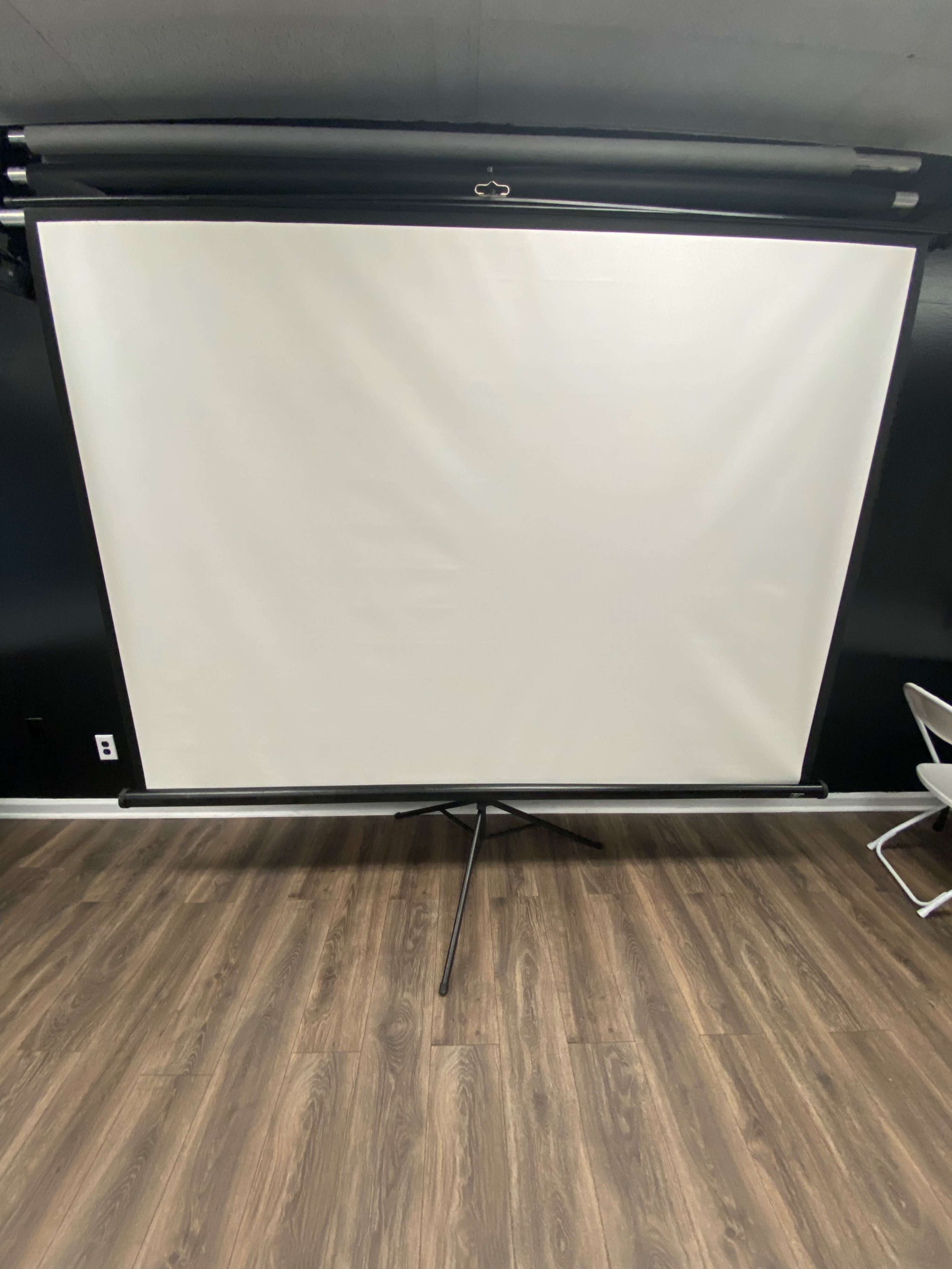 A projection screen is set up on a tripod in an office with a dark wall and wooden flooring.