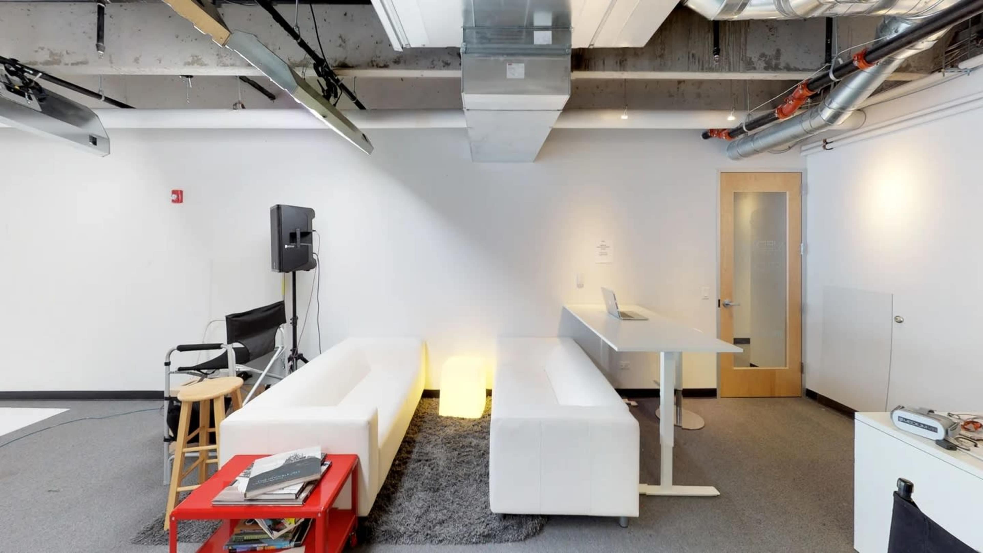 The image shows a modern, open interior space featuring a white sofa, a small table, and a few chairs, alongside some equipment and a desk.