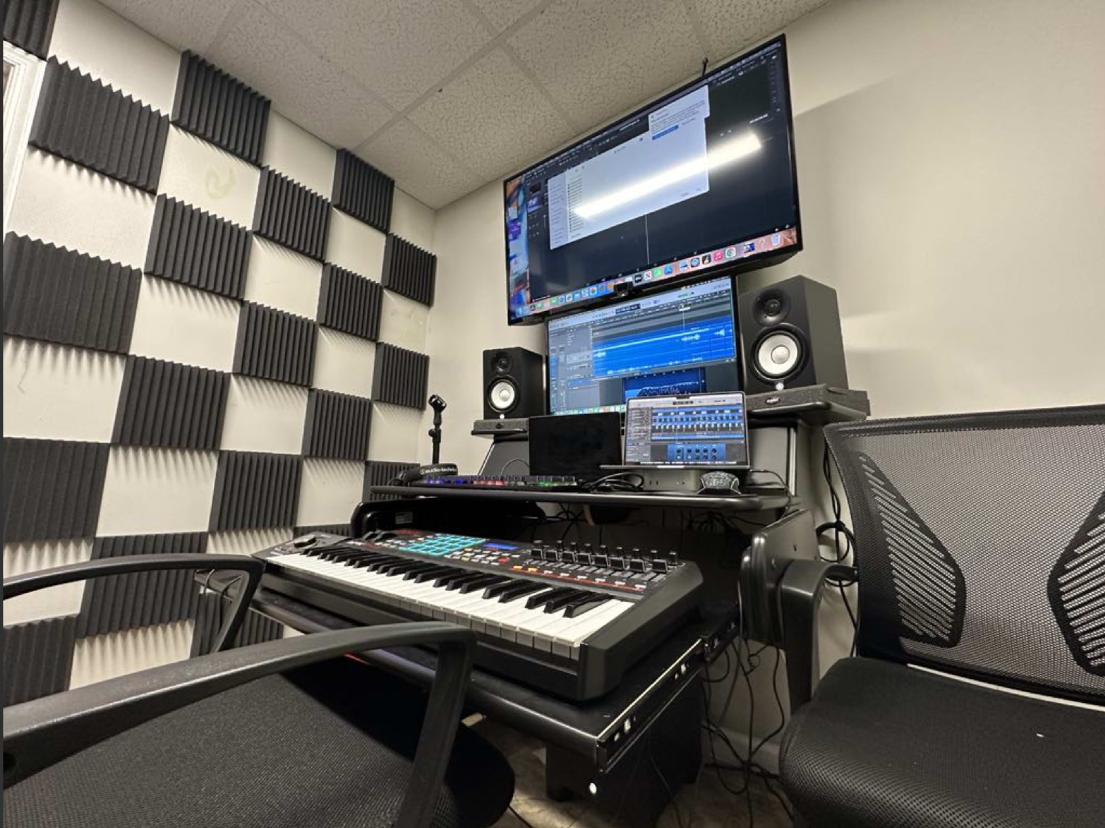 The image shows a music production studio featuring a keyboard, speakers, and a computer monitor displaying audio software, with soundproofing tiles on the walls.