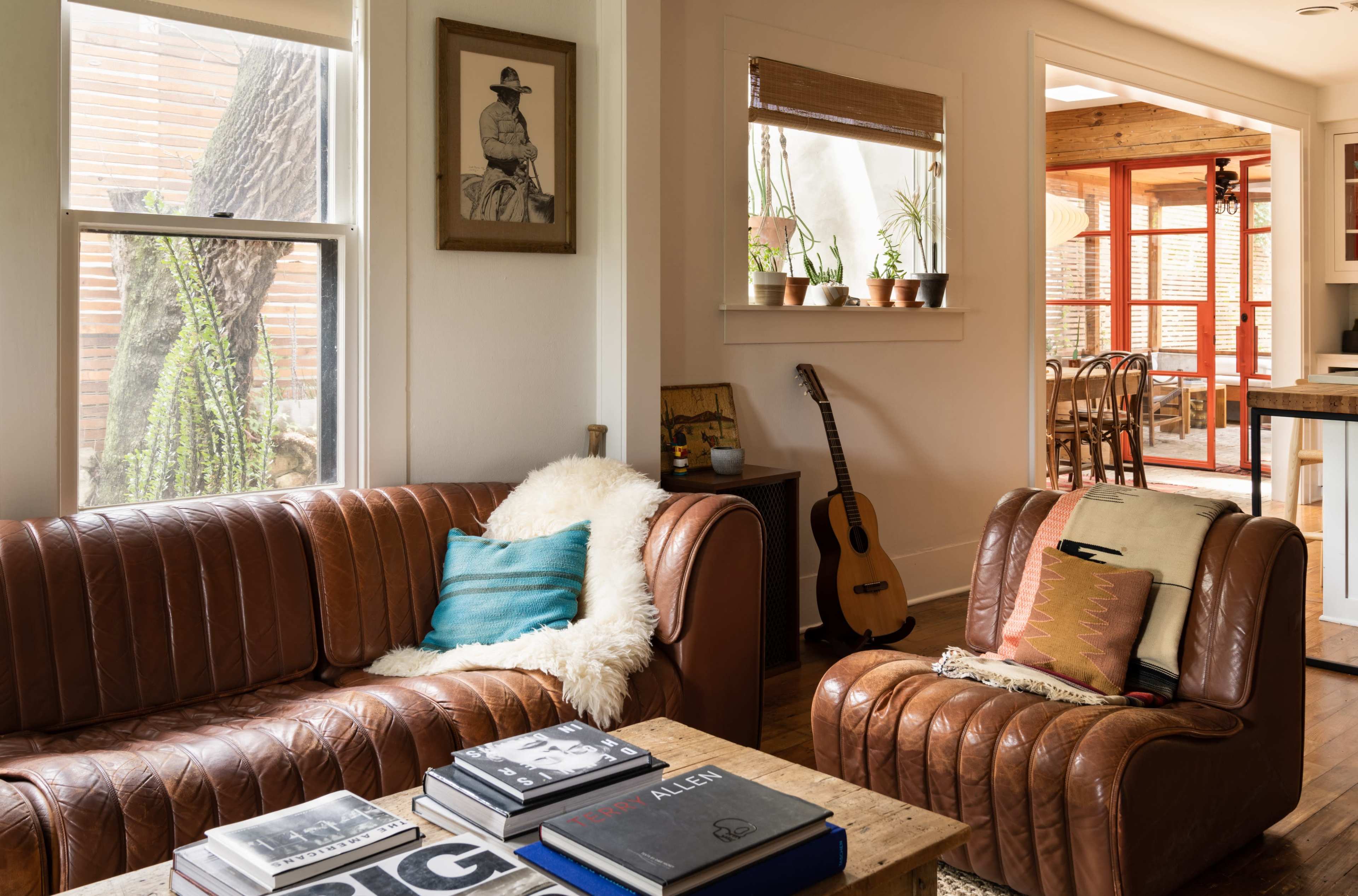The scene depicts a cozy living room with two leather sofas, a wooden coffee table stacked with books, and a guitar leaning against the wall, all illuminated by natural light coming through the windows.