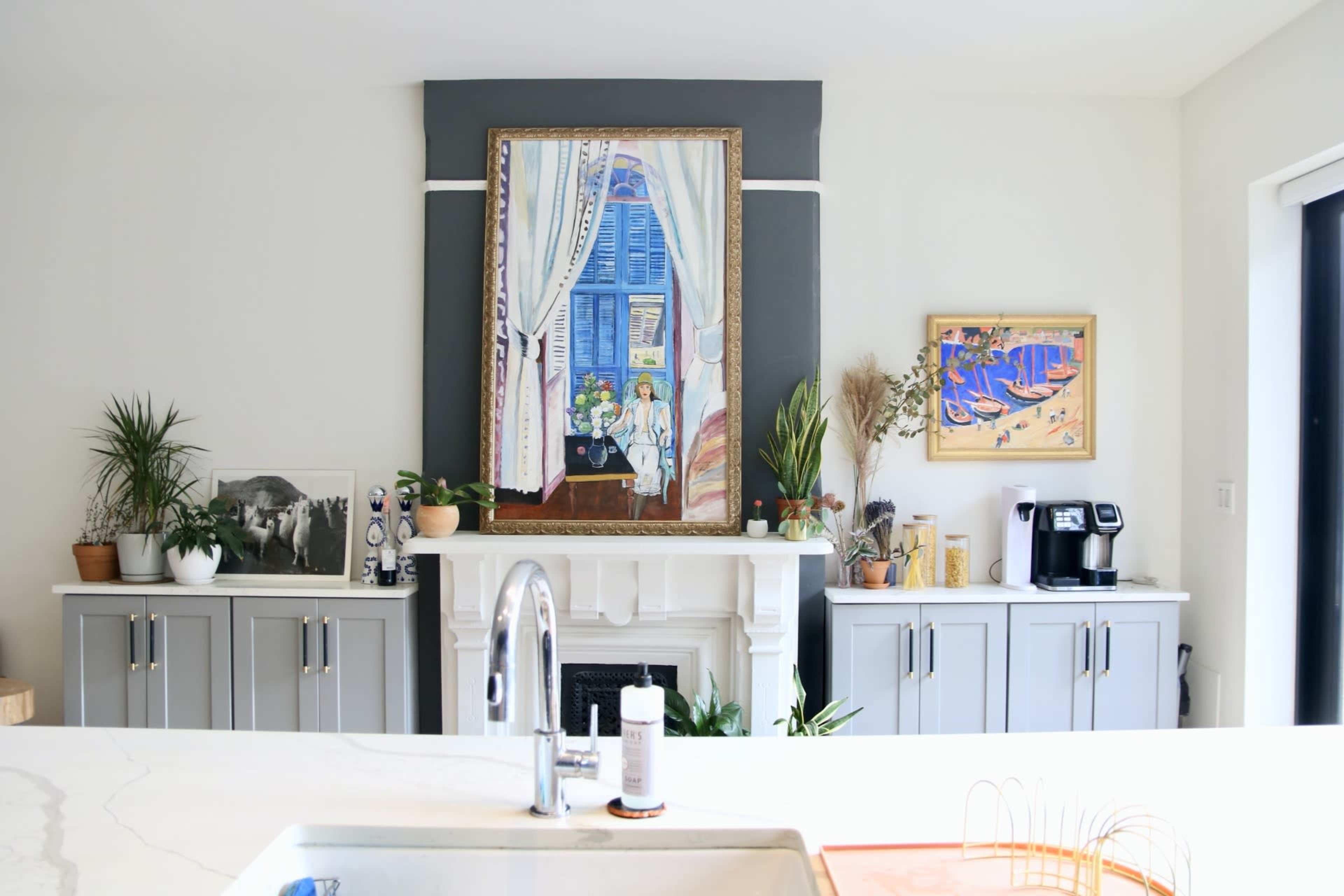 A kitchen with a central island, a large painting above a fireplace, and several potted plants on shelves and counters.