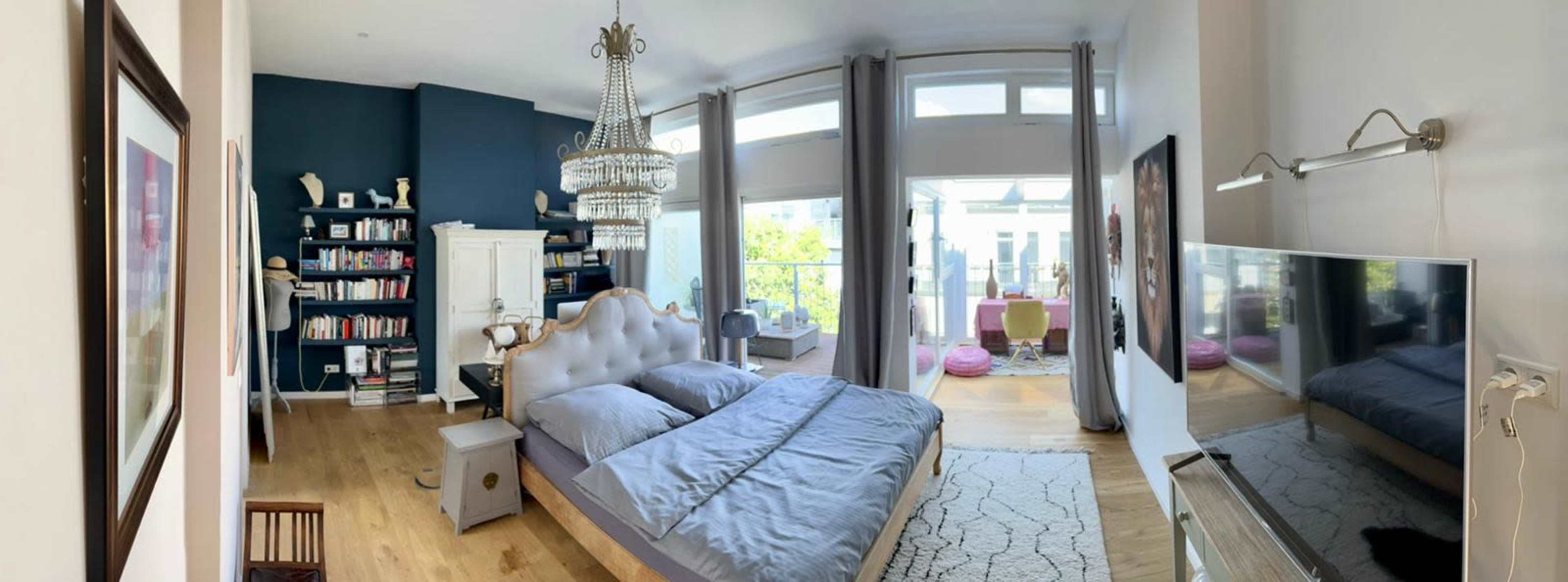 A modern bedroom with a large bed, a chandelier, bookshelves filled with books, and large windows that open to a balcony.