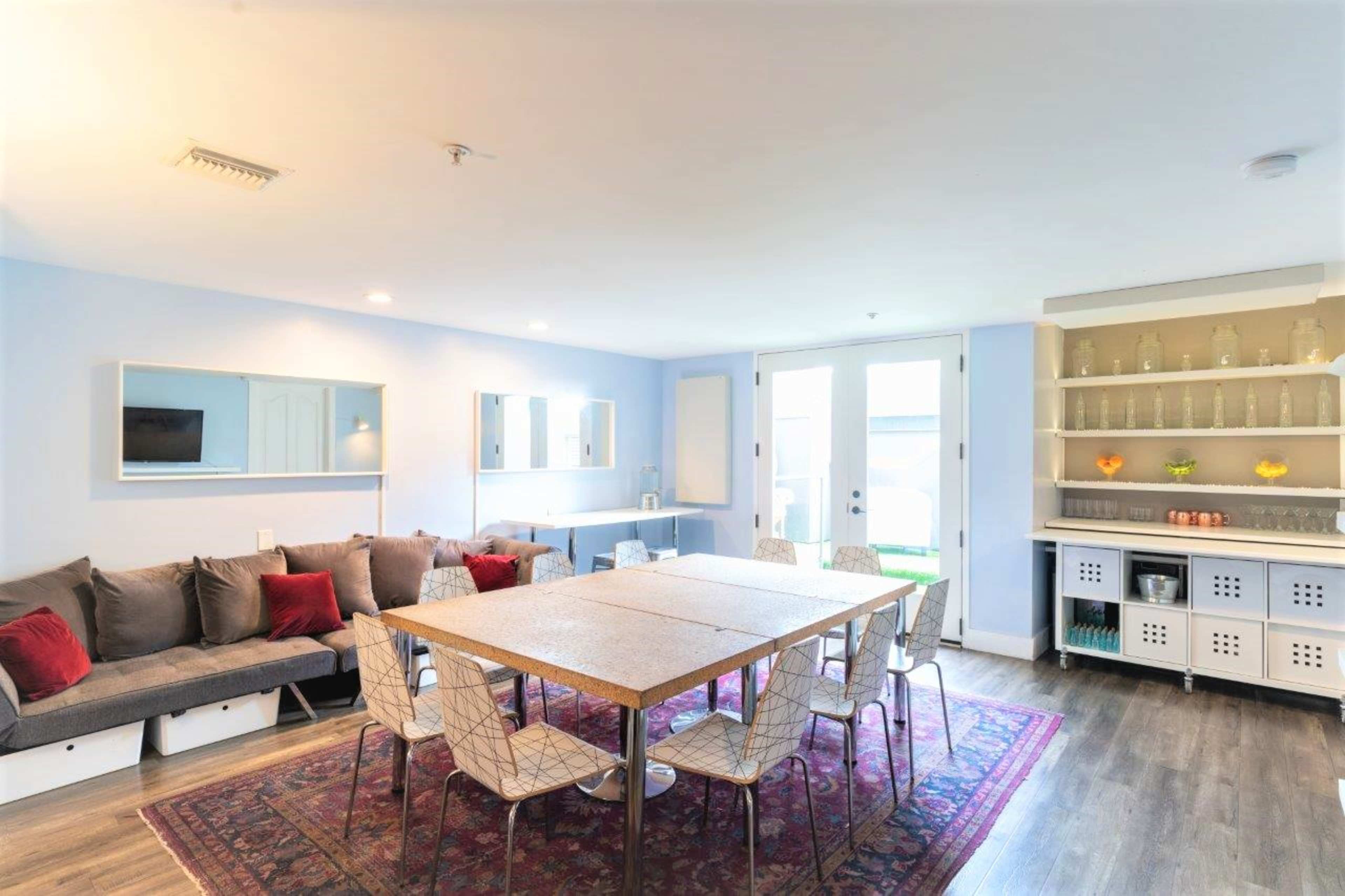 A dining area features a large table surrounded by chairs, with a sofa and a shelf filled with glassware in a well-lit, modern room.