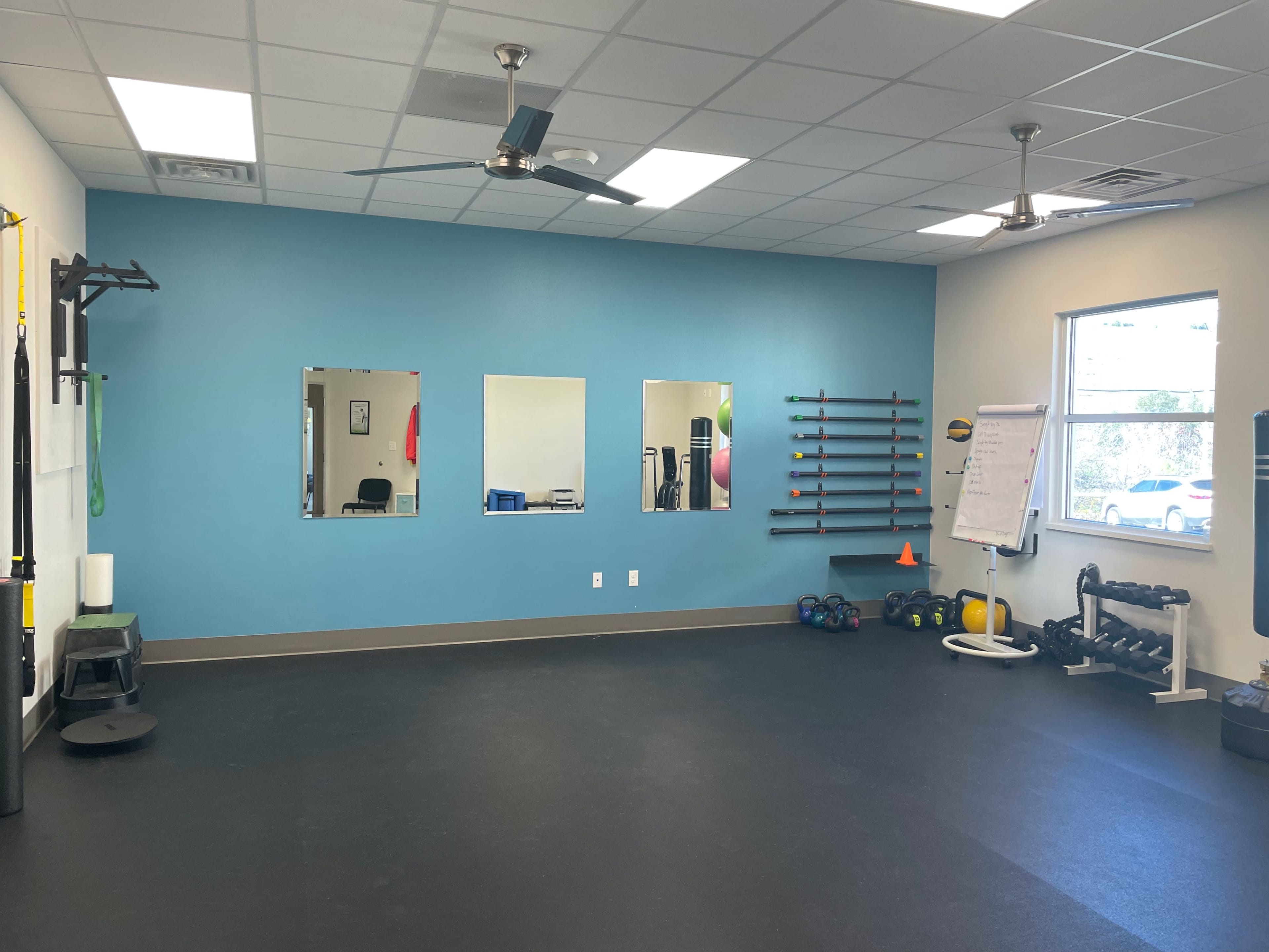 A well-lit gym interior featuring exercise equipment, three mirrors on a blue wall, and a black rubber floor.