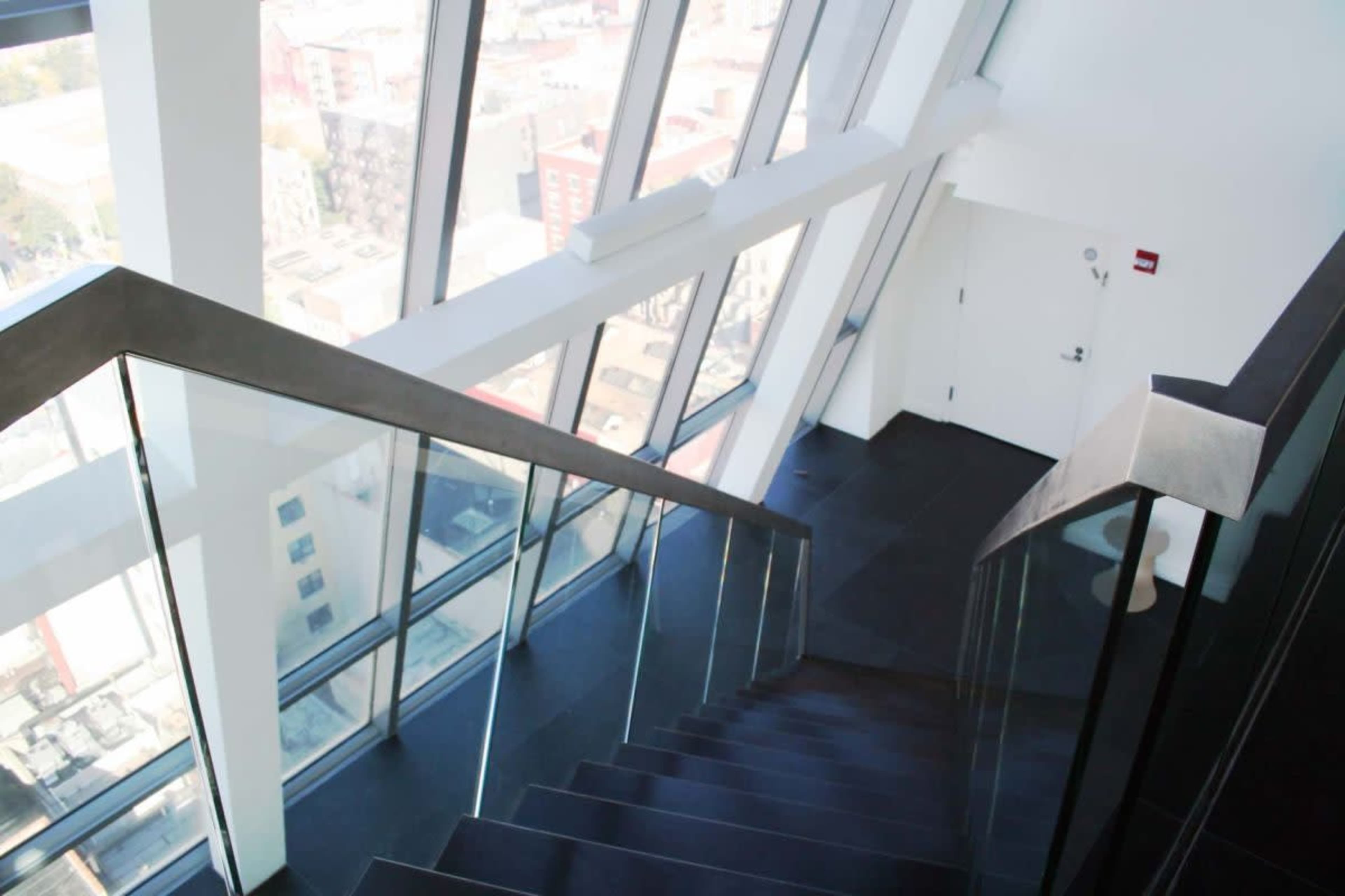 A staircase with glass railings descends from a modern building, leading to a door in the background with large windows revealing a city view.