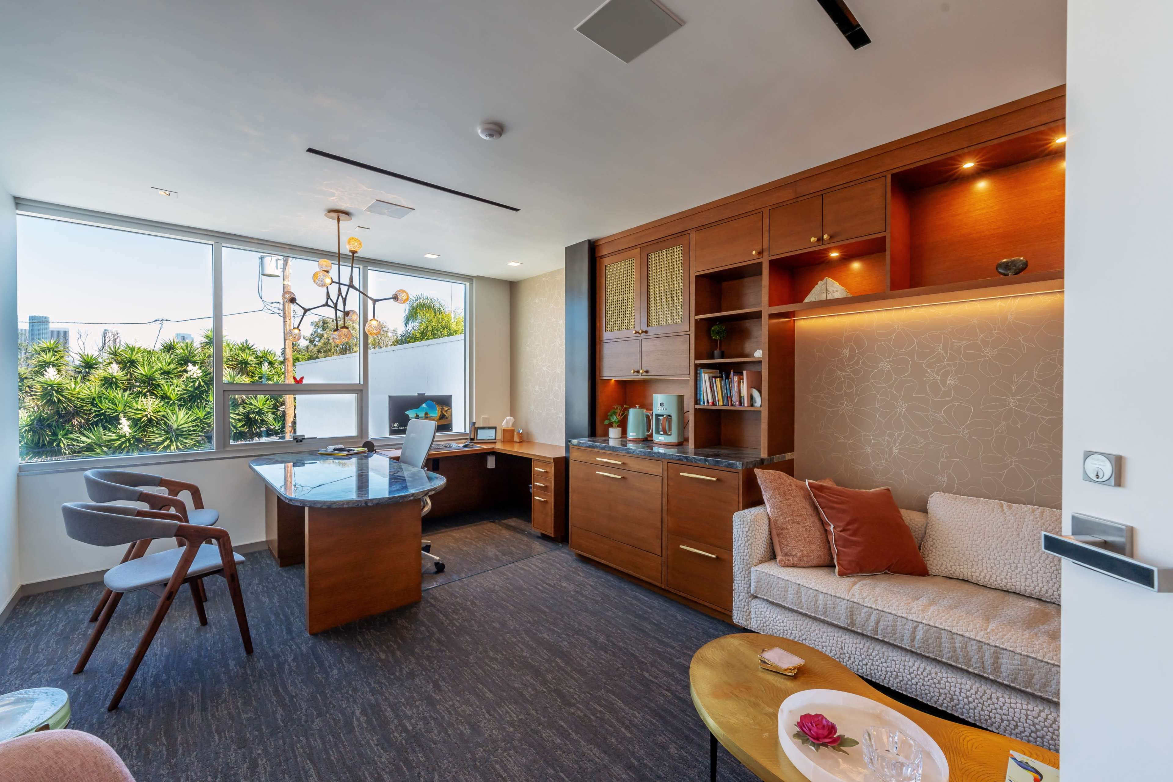 A modern office space featuring a desk with a computer, a circular glass table, and a comfortable sofa next to large windows.