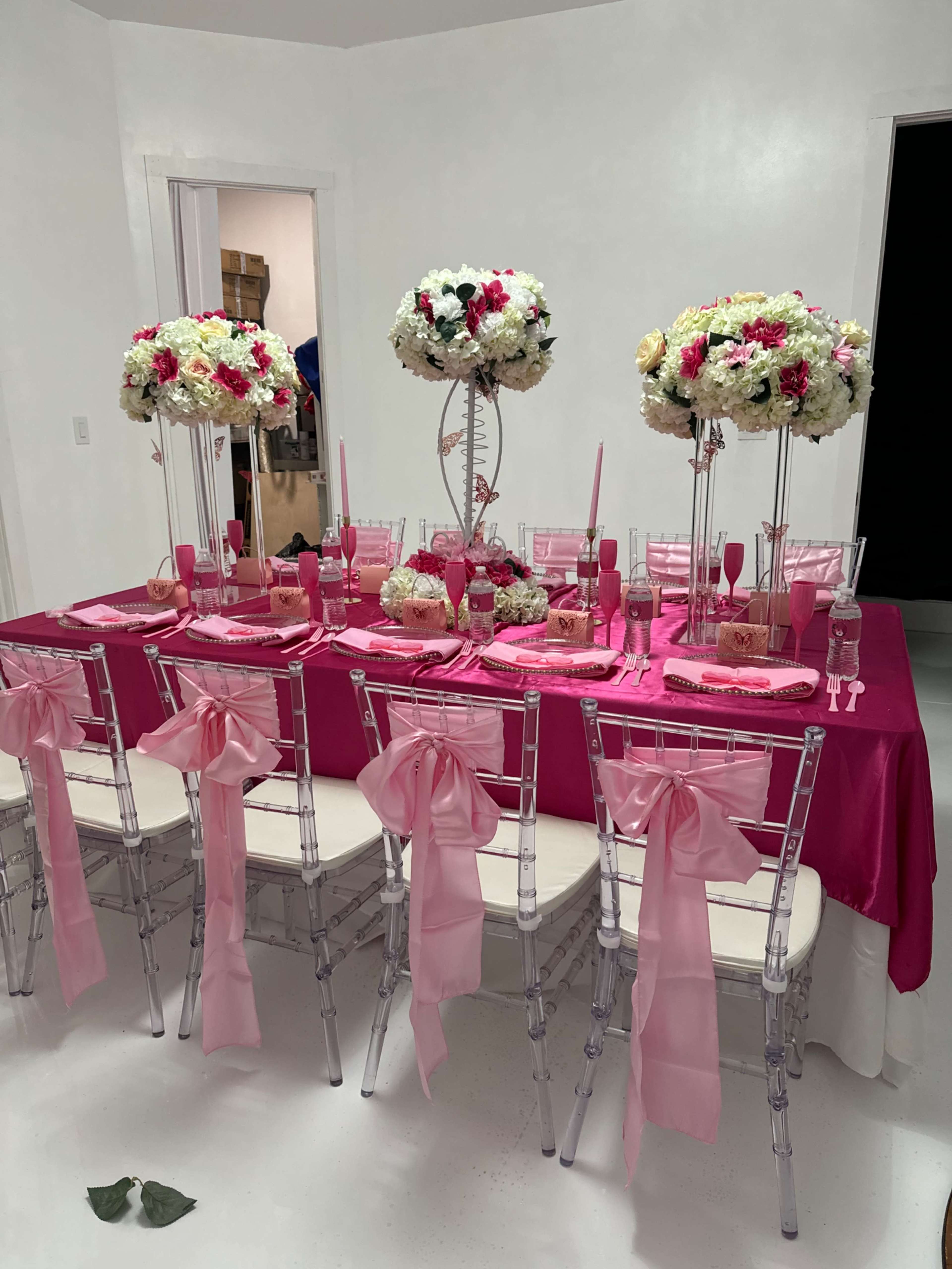 A large table decorated with pink and white floral arrangements sits in a bright room, surrounded by clear chairs with pink bows.