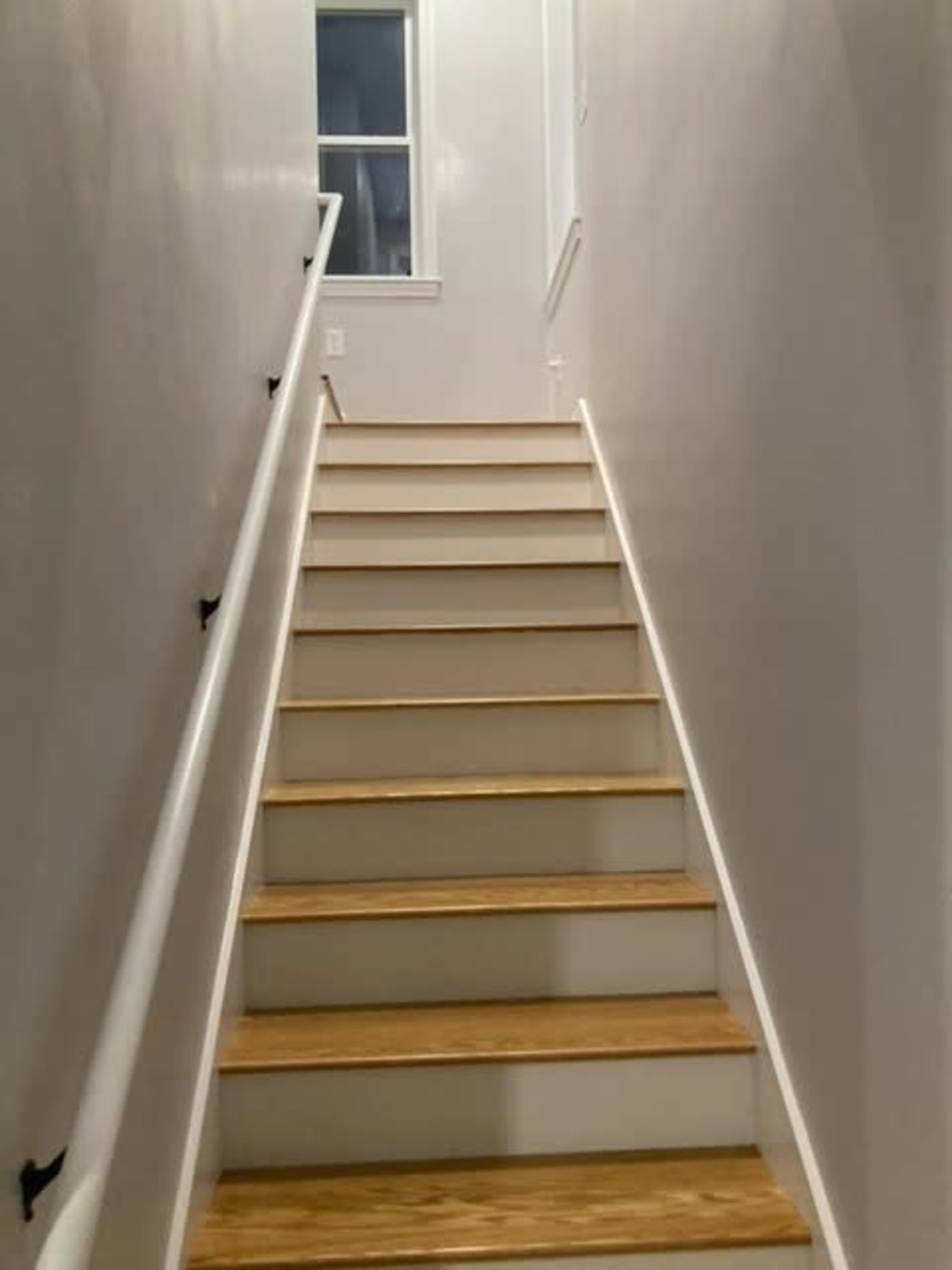 A wooden staircase leads upward in a well-lit hallway with white walls and a window at the top.