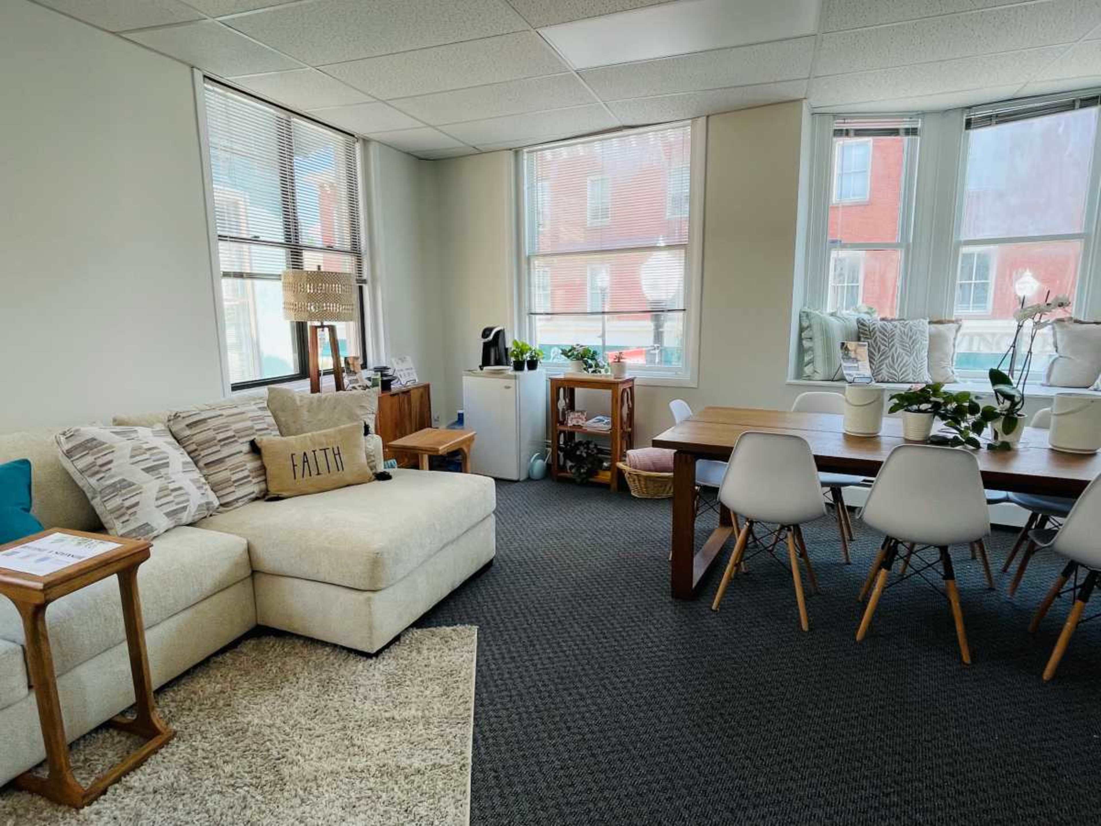 A light-filled room featuring a beige sectional sofa, a wooden dining table with white chairs, and potted plants on the windowsills.