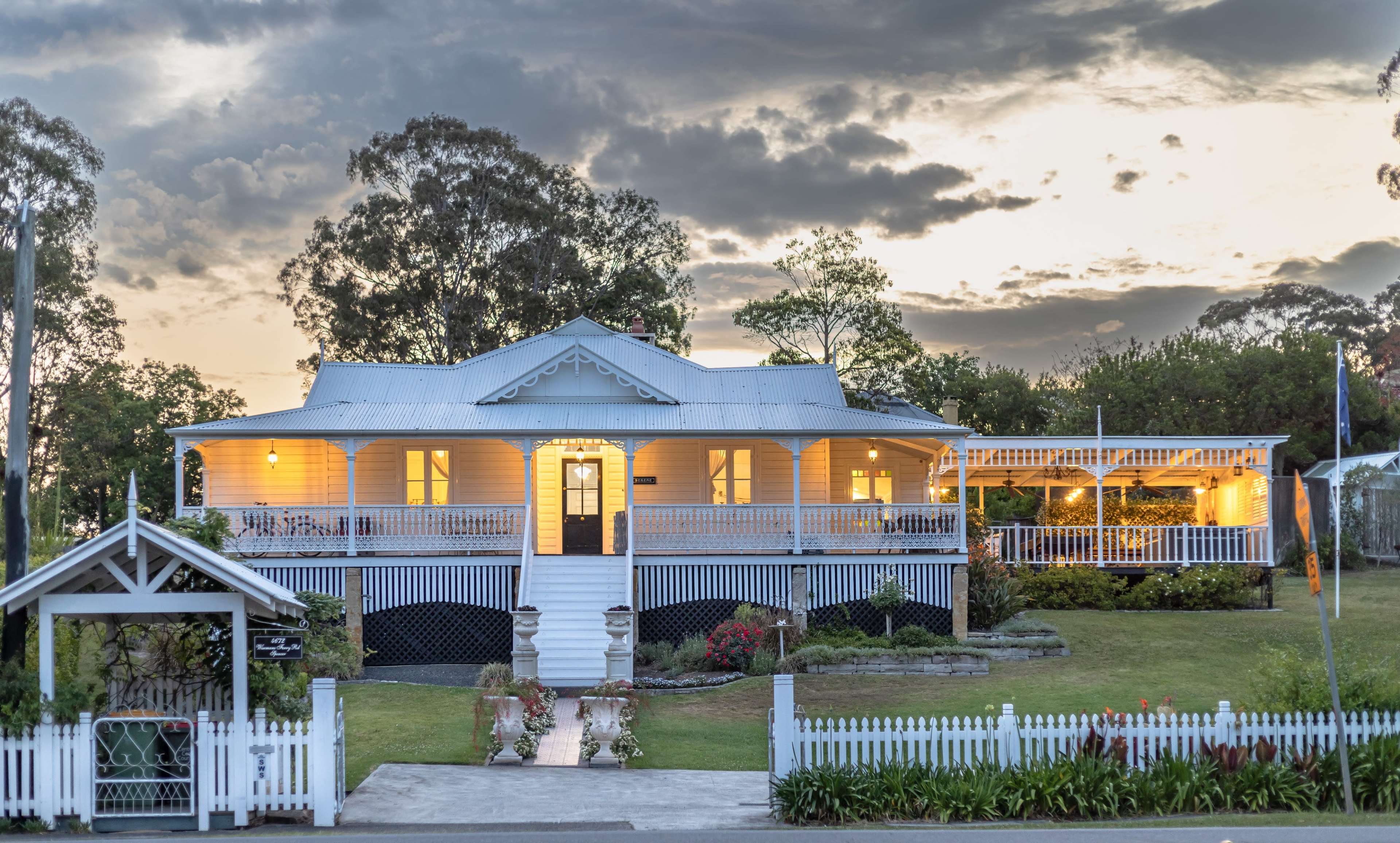 A spacious, white-clapboard house with a front porch and decorative railings sits amid landscaped gardens under a cloudy sky at dusk.