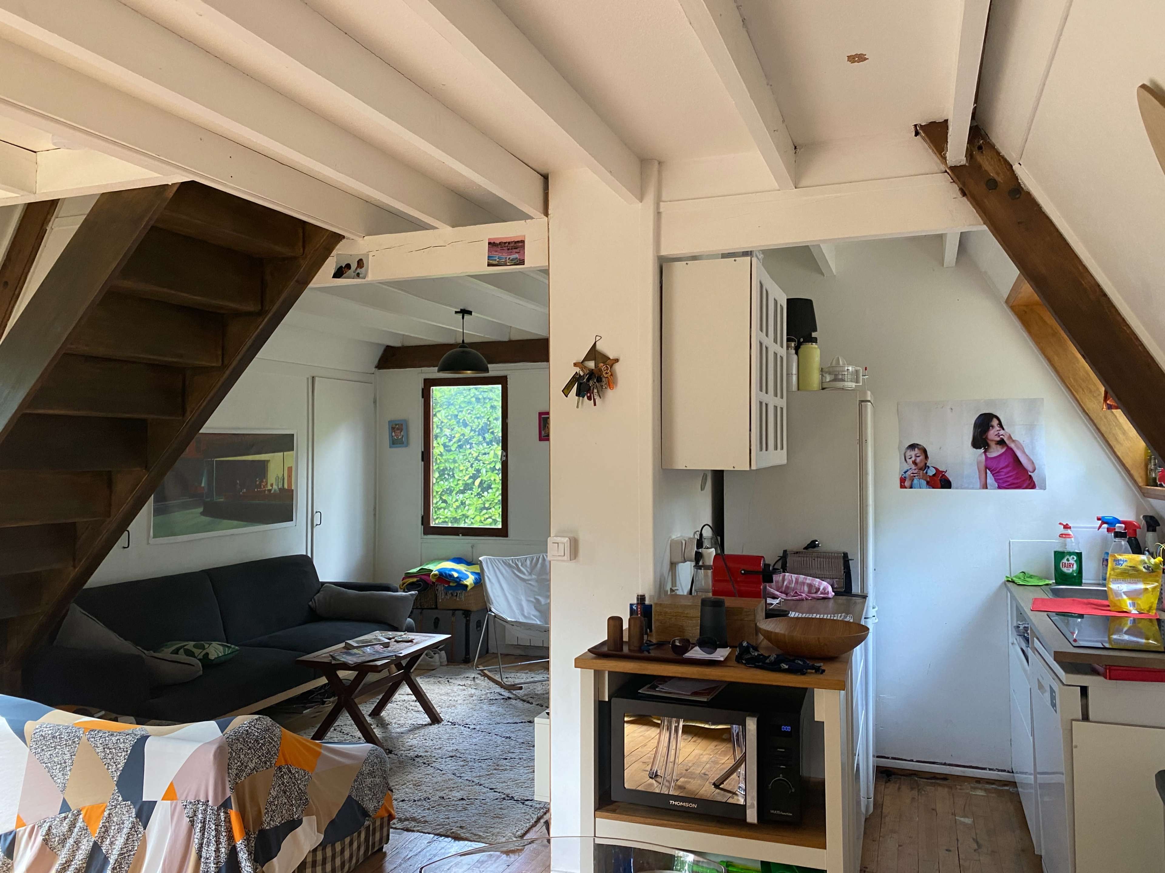 A compact living space features a staircase, a sofa, a kitchen area, and a window with outdoor visibility.