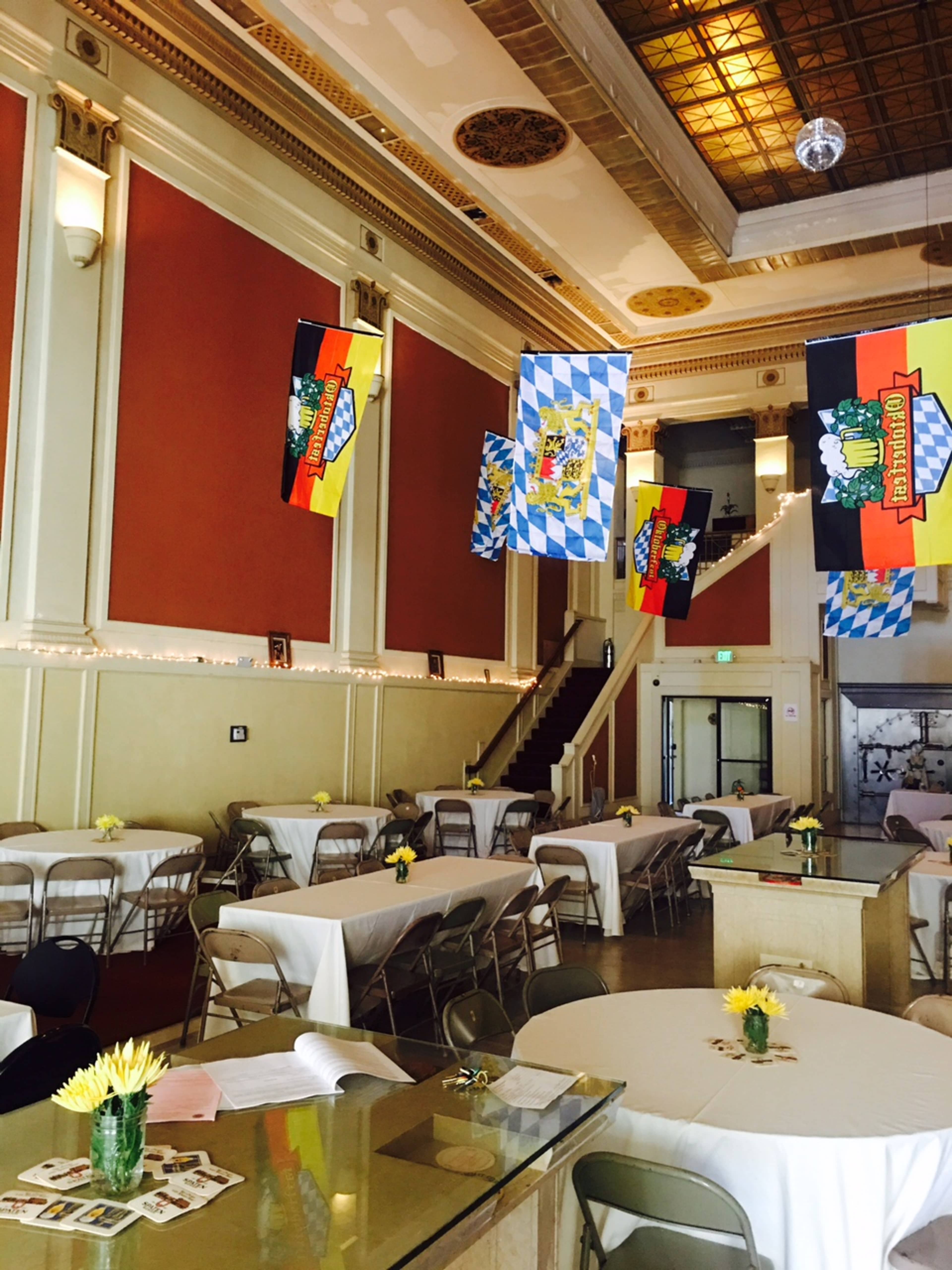 A spacious event hall features tables covered with white linens, adorned with sunflowers, and decorated with colorful banners hanging from the ceiling.