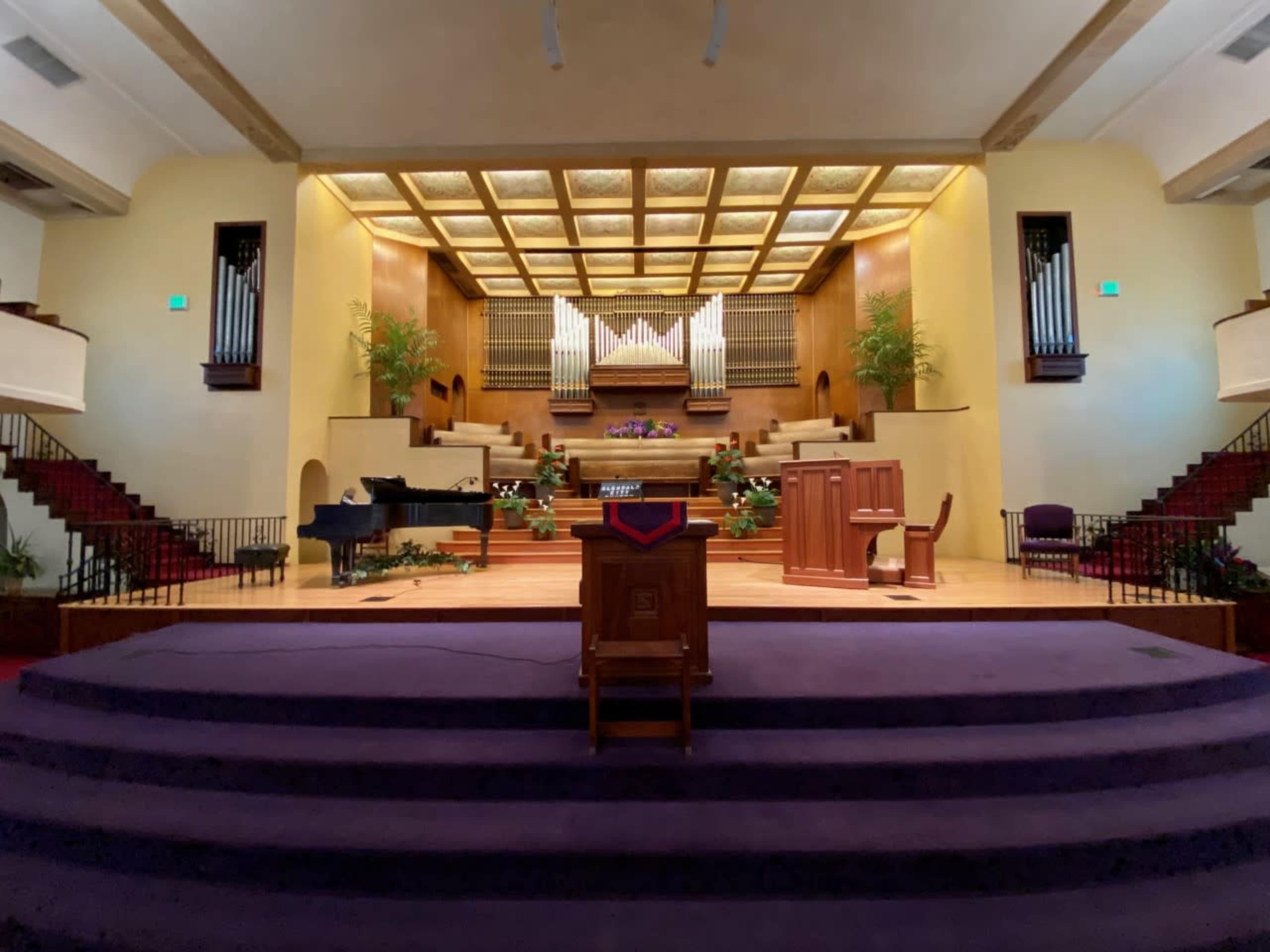 The interior of a church sanctuary with a raised pulpit, a grand piano, and an organ setup, surrounded by wooden paneling and green plants.
