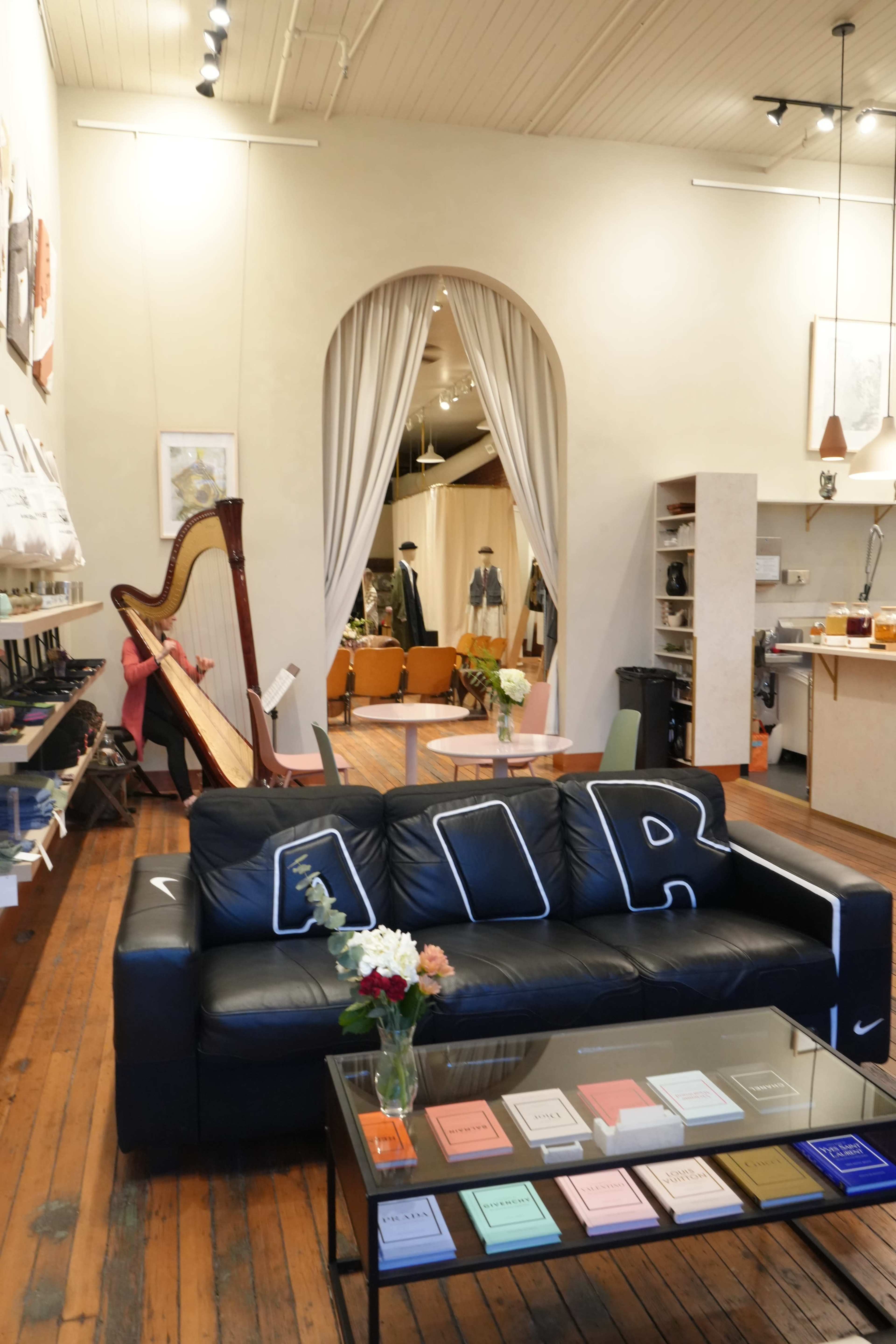 The image shows a stylish interior of a boutique or store featuring a black sofa with decorative pillows, a harp in the background, and various products displayed on shelves and tables.
