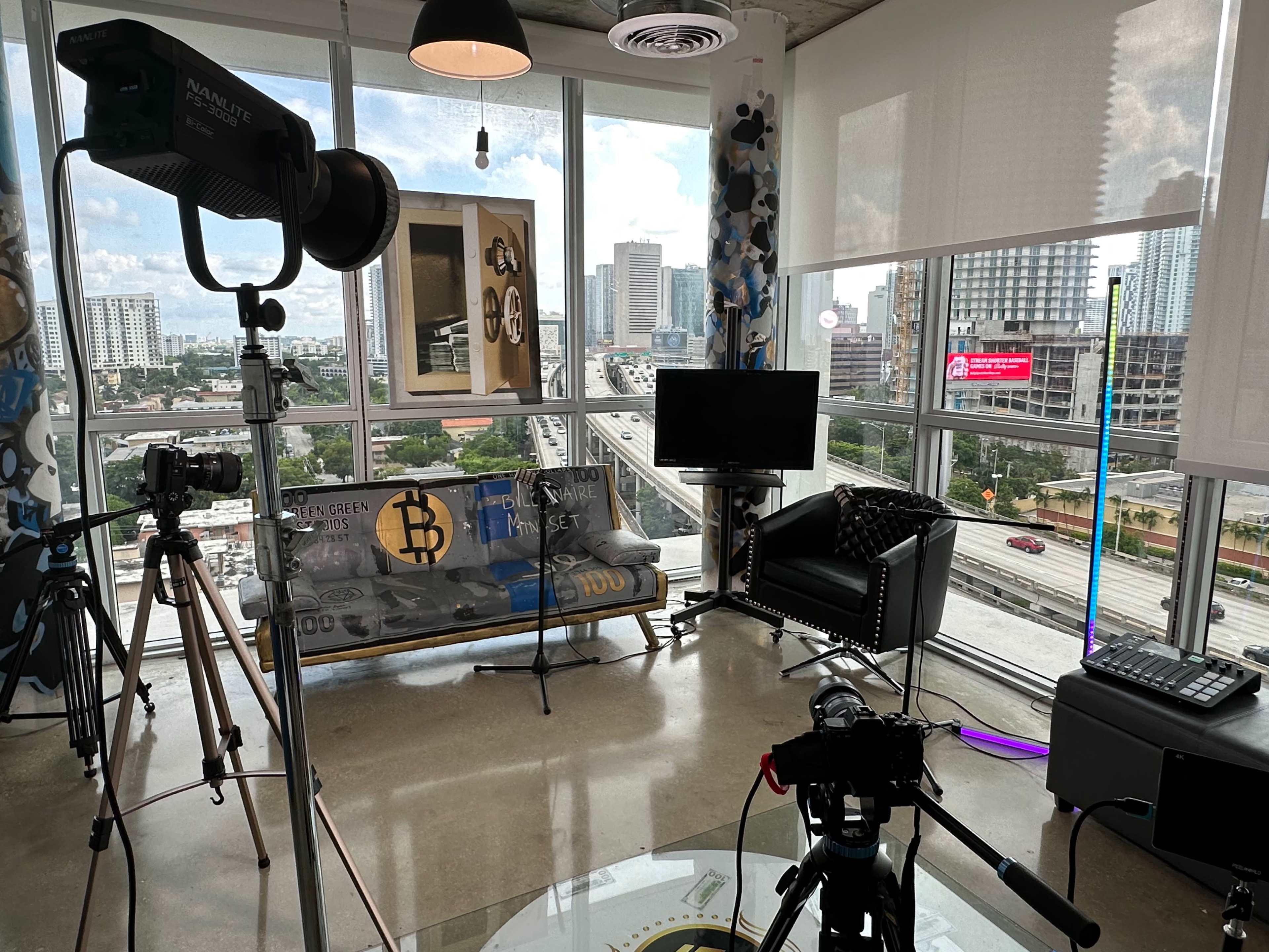 A modern studio space features a couch, television, and camera equipment, with large windows overlooking a city street.