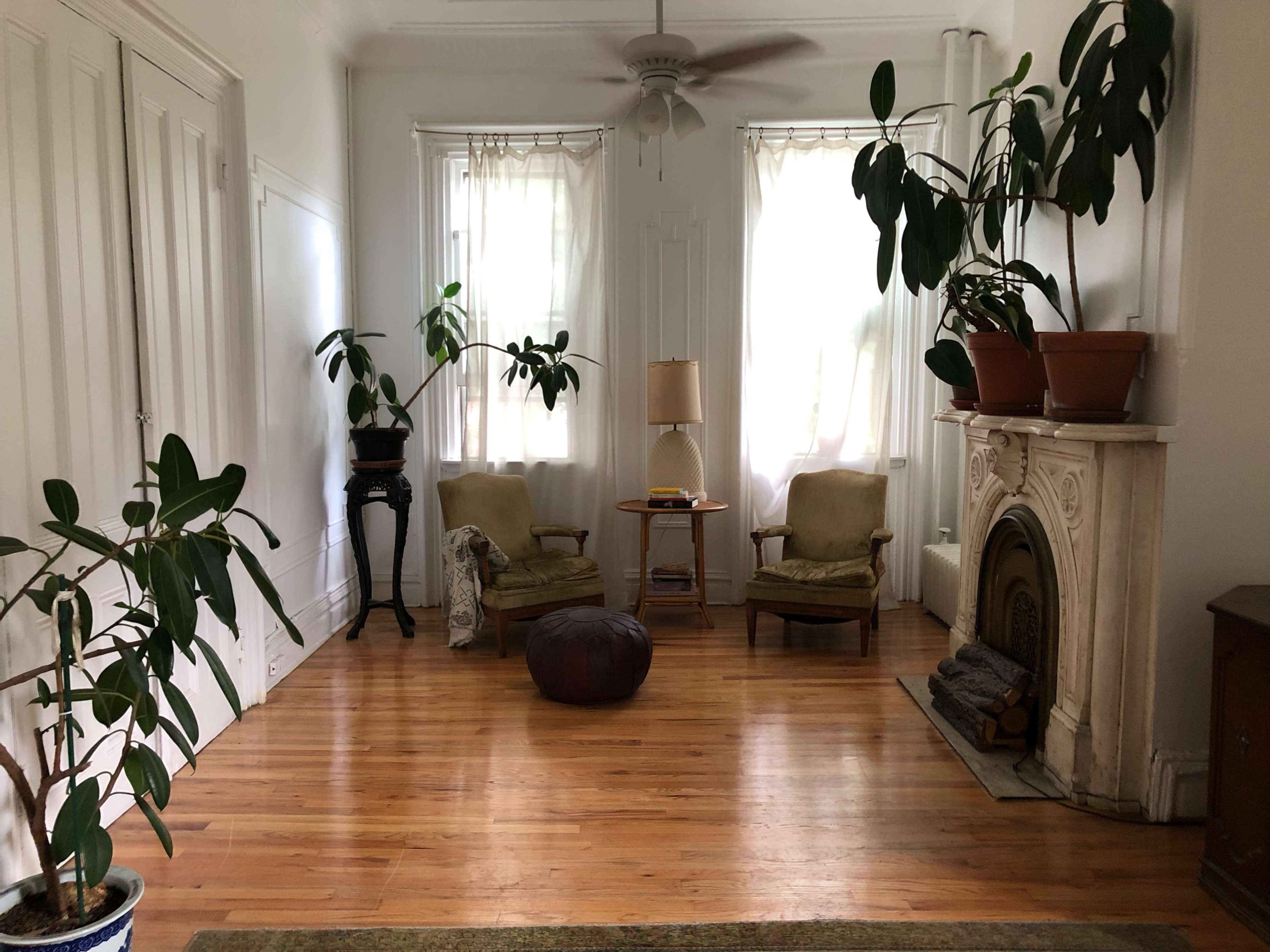 A spacious living room with two chairs, a small table, and several potted plants near a decorative fireplace.