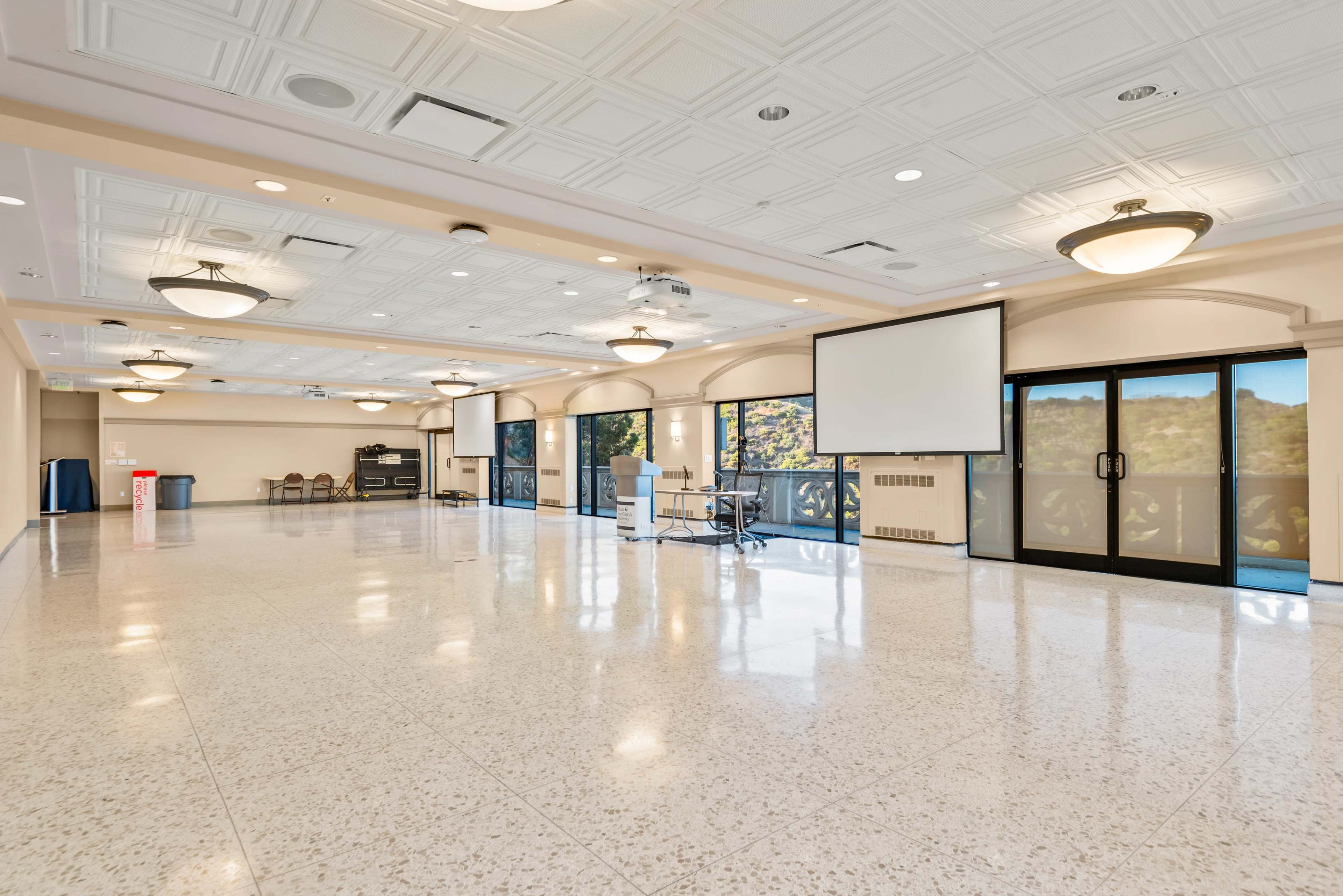 The image depicts a spacious, empty indoor hall with large windows, a podium, and two projection screens.