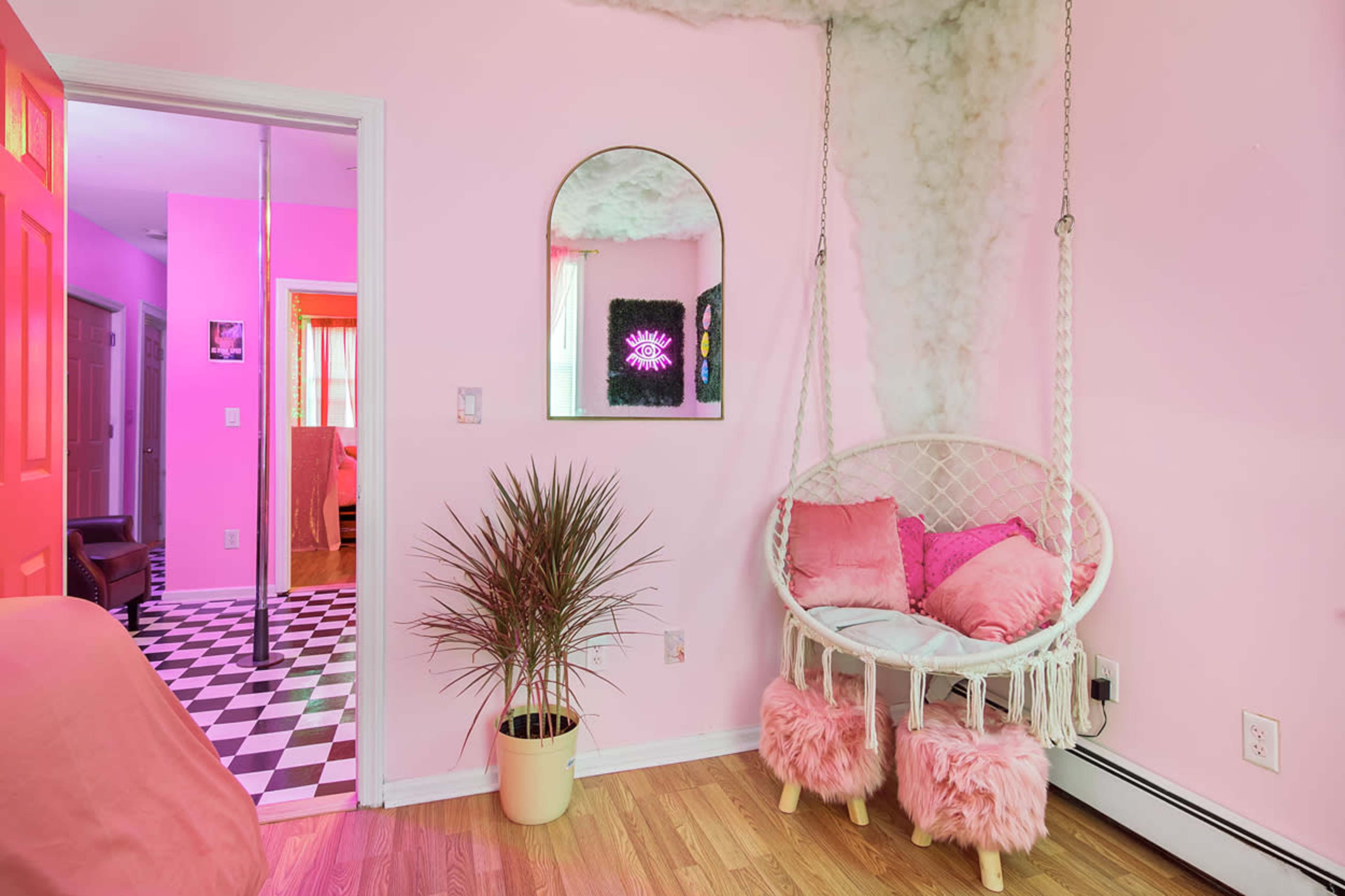 A pink-themed room featuring a hanging chair with plush cushions, a potted plant, and a mirror, with a checkered floor visible in the background.