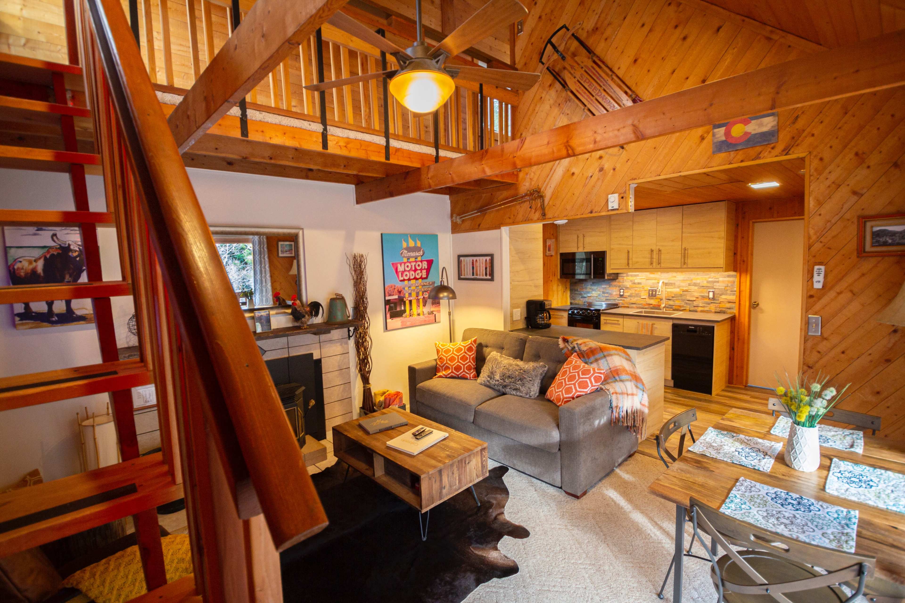 A cozy living space with wooden beams, a staircase leading to a loft, a fireplace, a sofa, and a kitchen area.
