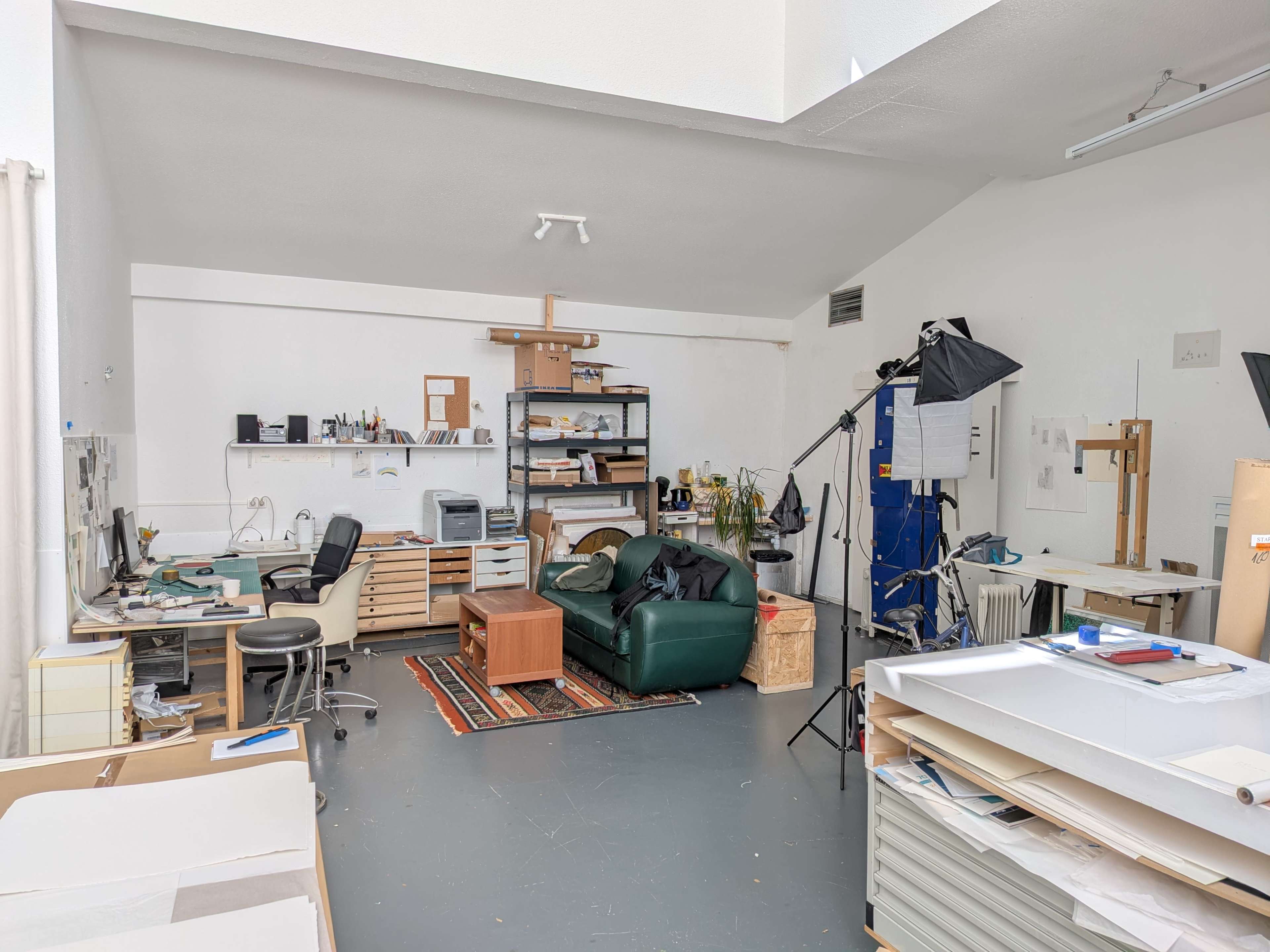 The image shows an art studio with a green sofa, work tables, shelves filled with supplies, and equipment for photography.