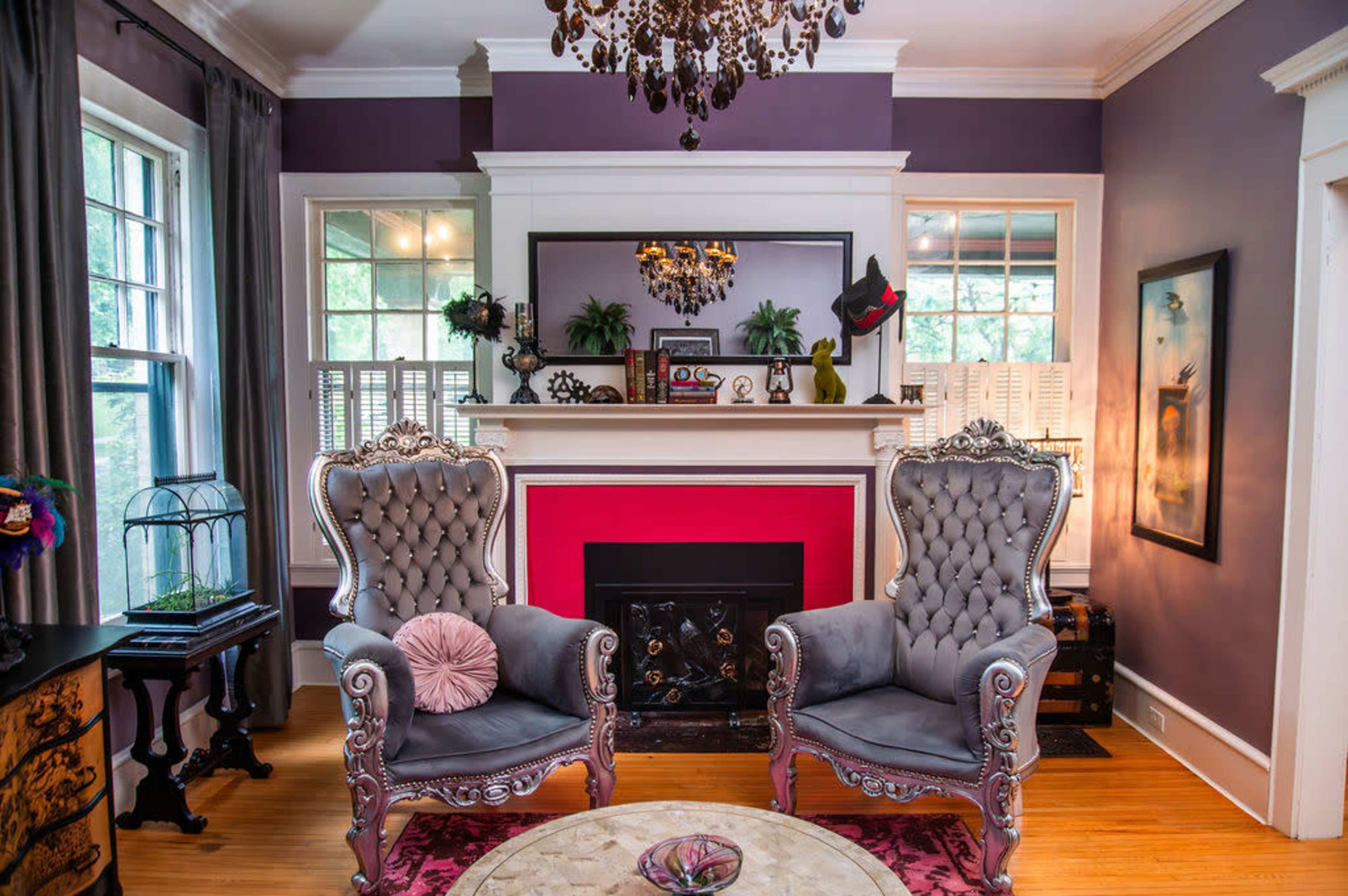 The image depicts a stylish living room with two ornate gray armchairs facing a bright red fireplace, surrounded by purple walls and elegant decor.